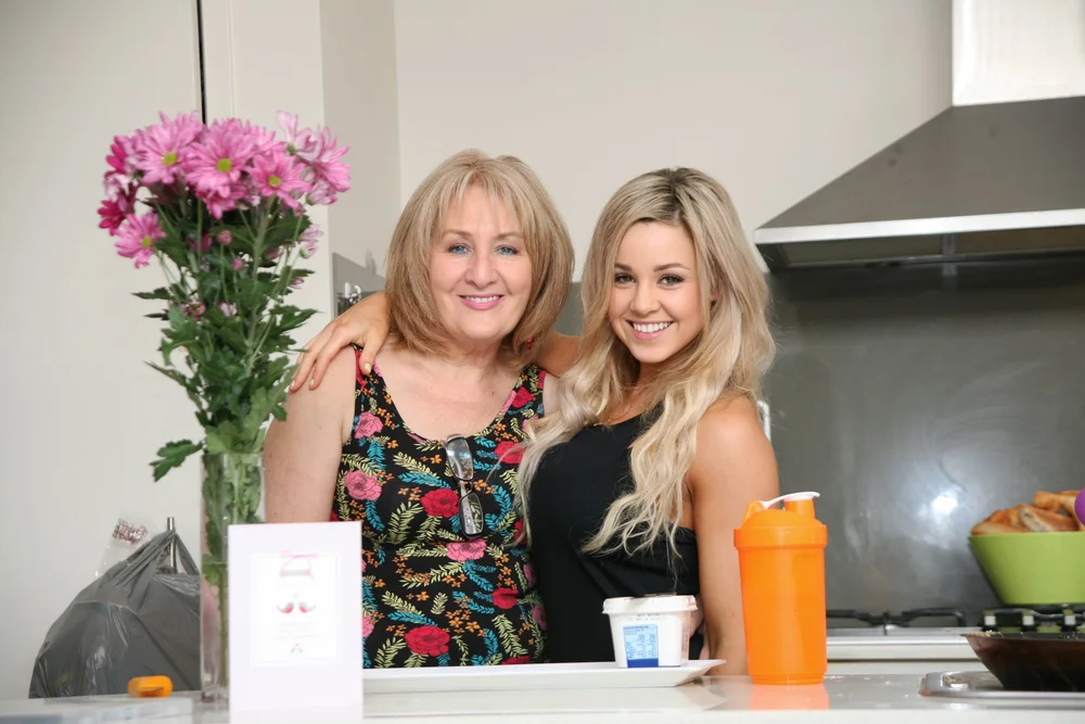 Two women smiling in a kitchen, one with blonde hair wearing a black top, the other with a floral dress. Visible items include pink flowers, an orange bottle, and a card.