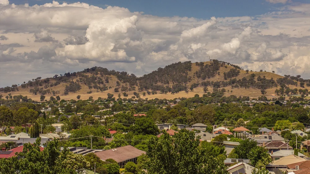Scenic view of a small town with houses and lush greenery, set against rolling hills under a cloudy sky.