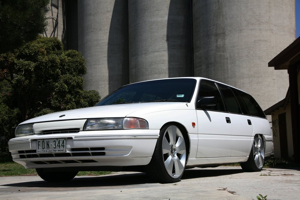 White station wagon parked in front of large concrete silos, with custom rims, green foliage, and blue sky.