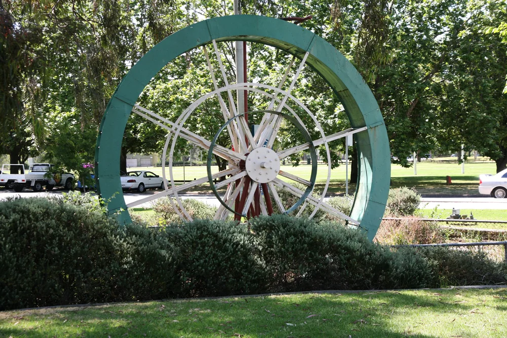 Large green decorative wheel structure in a park with trees and cars in the background.