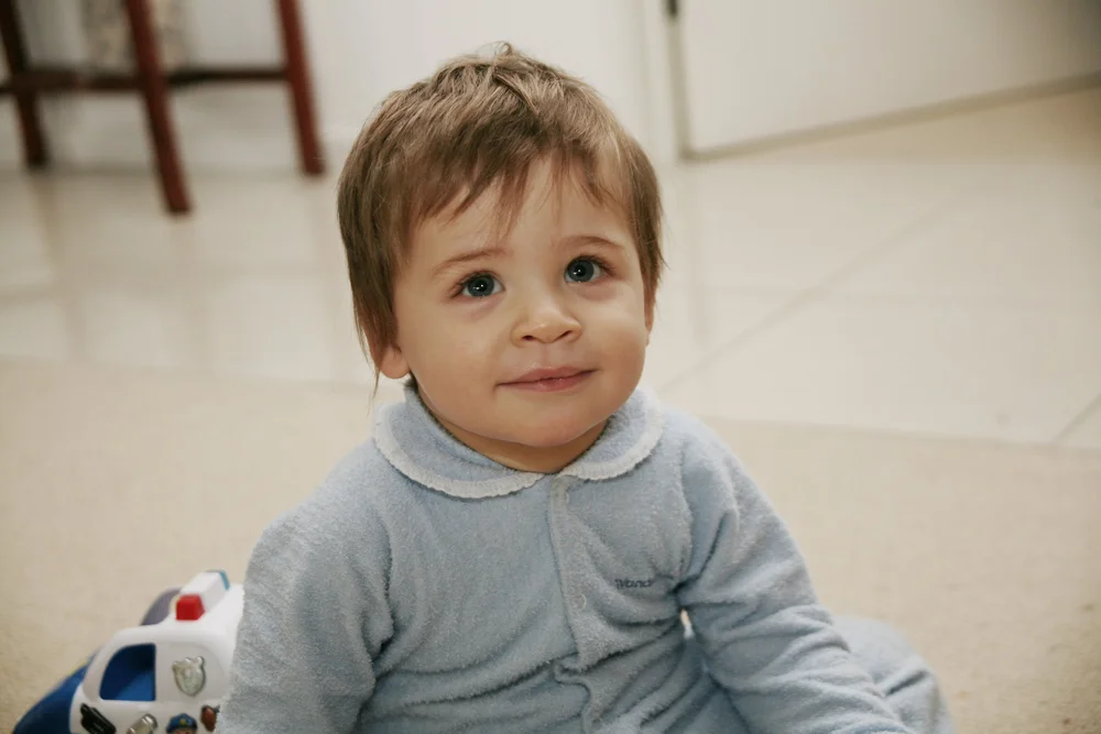 Toddler sitting on floor wearing light blue pajamas and a toy police car visible in background.
