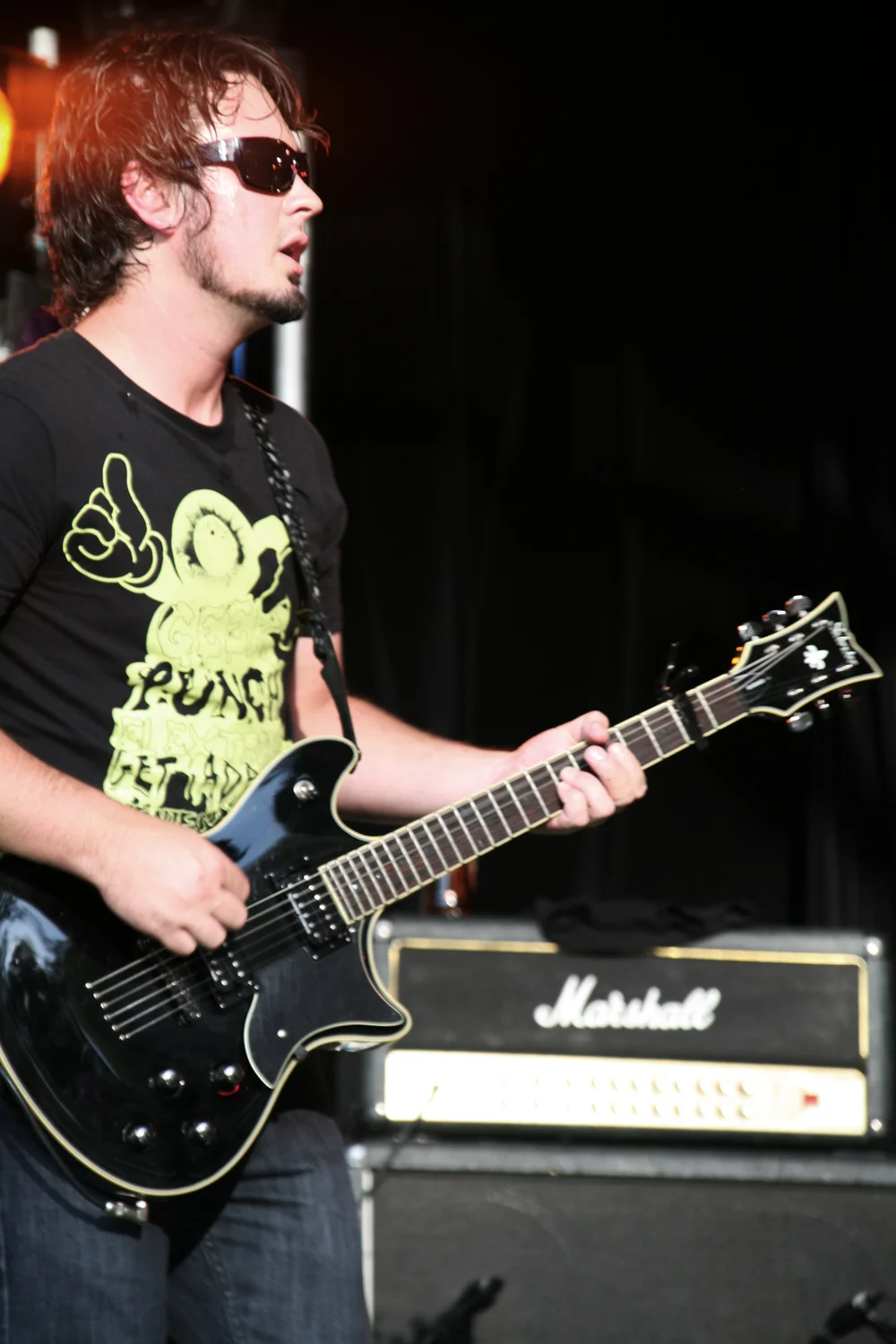 Guitarist playing electric guitar on stage with amplifier in background, wearing sunglasses and a graphic t-shirt.