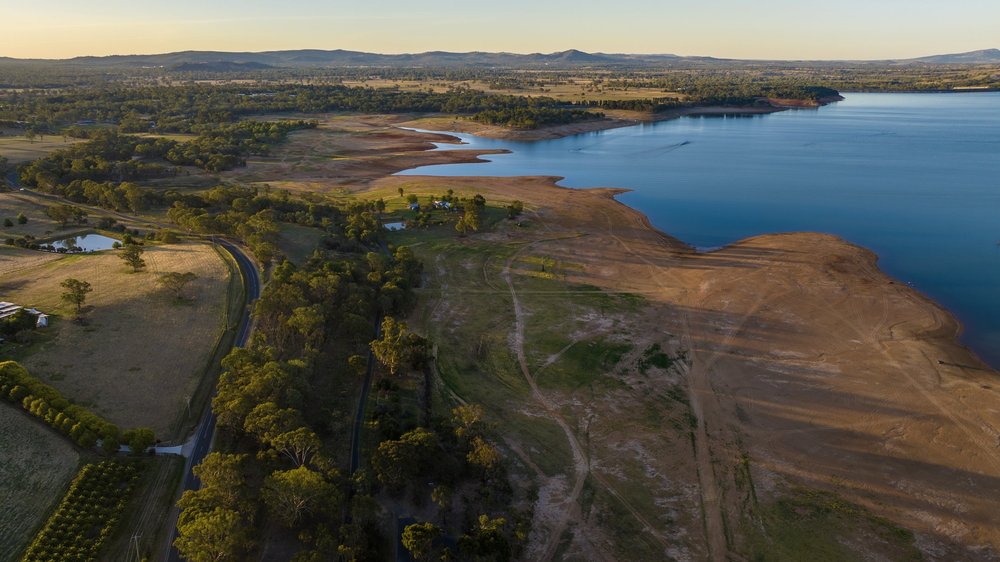 Aerial view of a lake with dry shoreline, surrounded by green trees and grasslands, with a road and farmland visible.