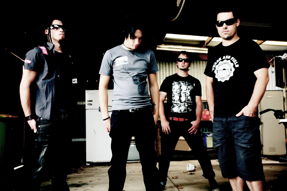 Four men in casual clothing and sunglasses in an indoor setting, with a fridge and tools visible in the background.