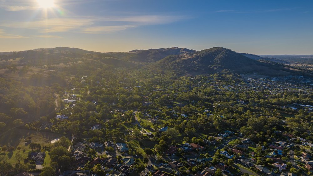 Aerial view of a suburban landscape with houses, trees, and hills under a bright sun.