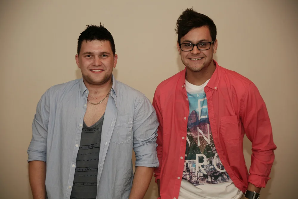 Two men smiling, wearing casual shirts and standing in front of a plain background.