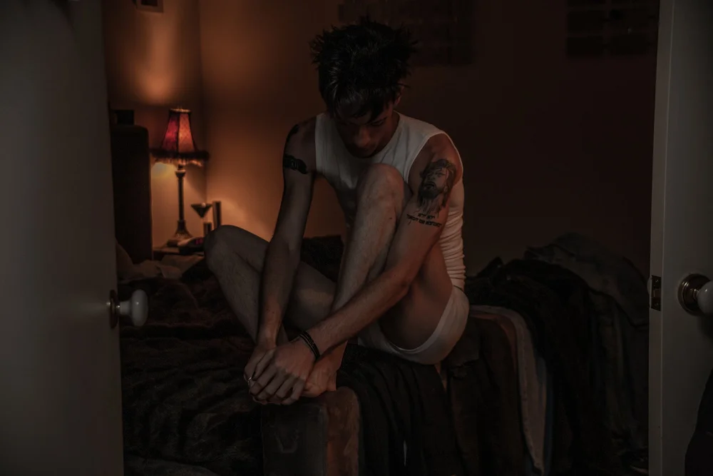Person sitting on bed in dimly lit room, wearing a white tank top and shorts, with a tattoo on their arm. A lamp is on a bedside table in the background.