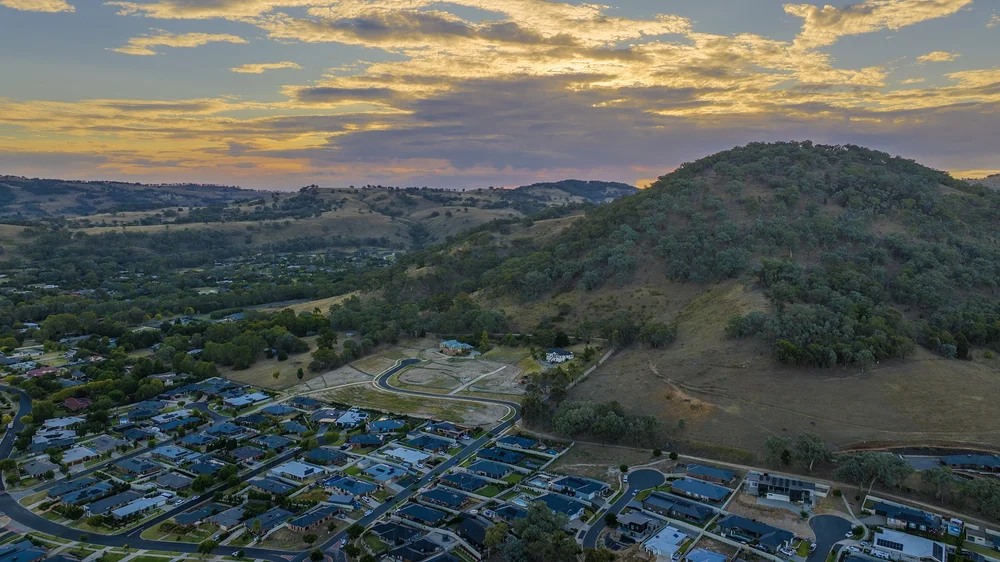 Aerial view of residential housing and a green hillside under a cloudy sky at sunset.