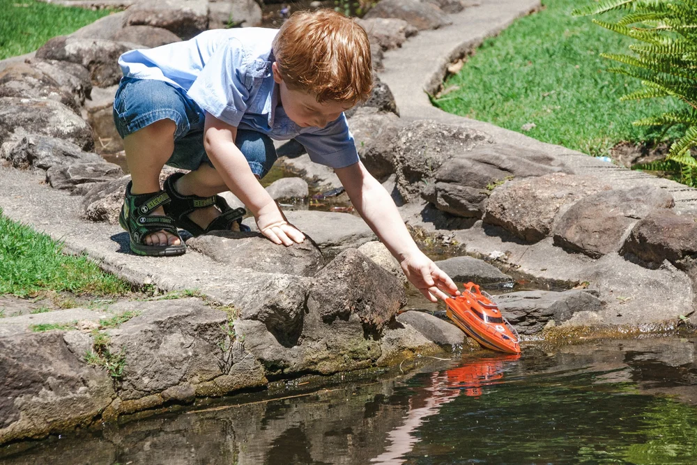 Child playing with toy boat near a water feature in a park.