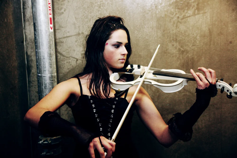 Woman playing a white electric violin against a textured wall background.