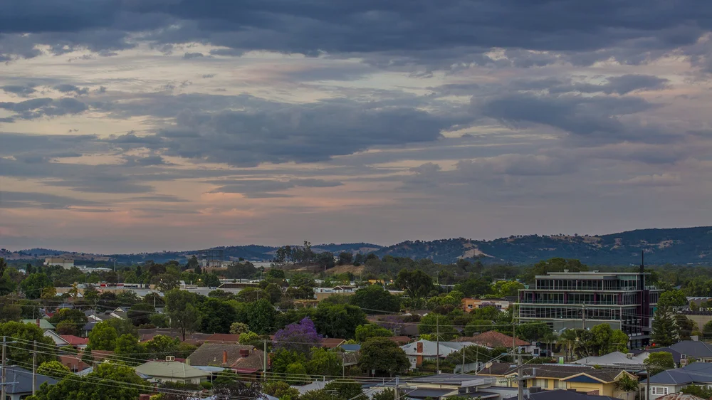 Suburban landscape with residential houses, trees, and distant hills under a cloudy sky at dusk.