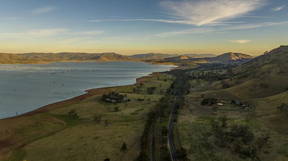 Aerial view of a wide landscape with a lake, rolling hills, and scattered trees under a blue sky.
