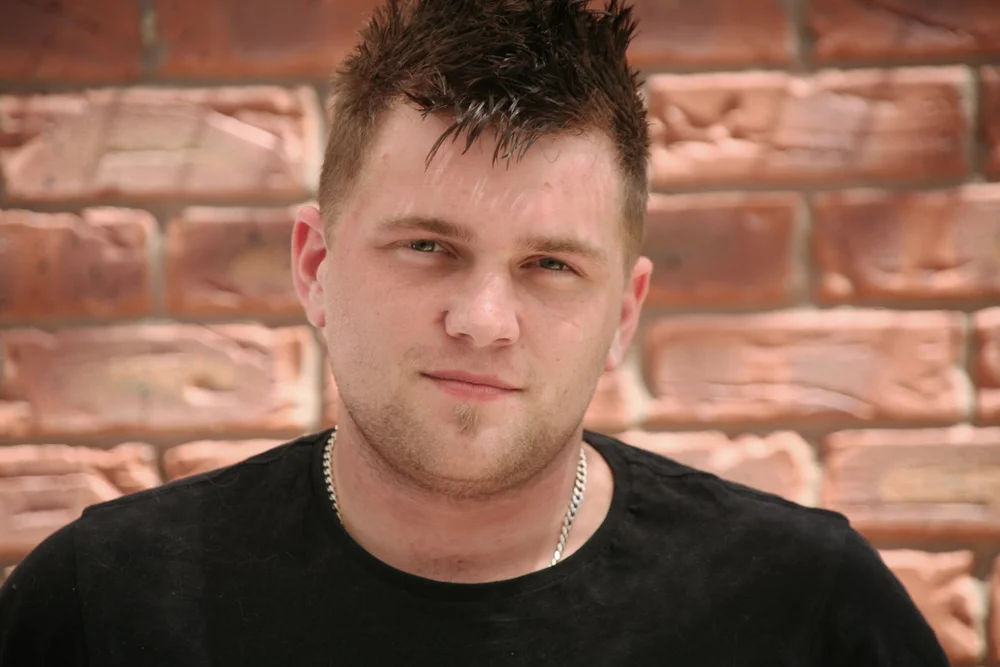 Man with spiked hair standing against a brick wall, wearing a black shirt and chain necklace.