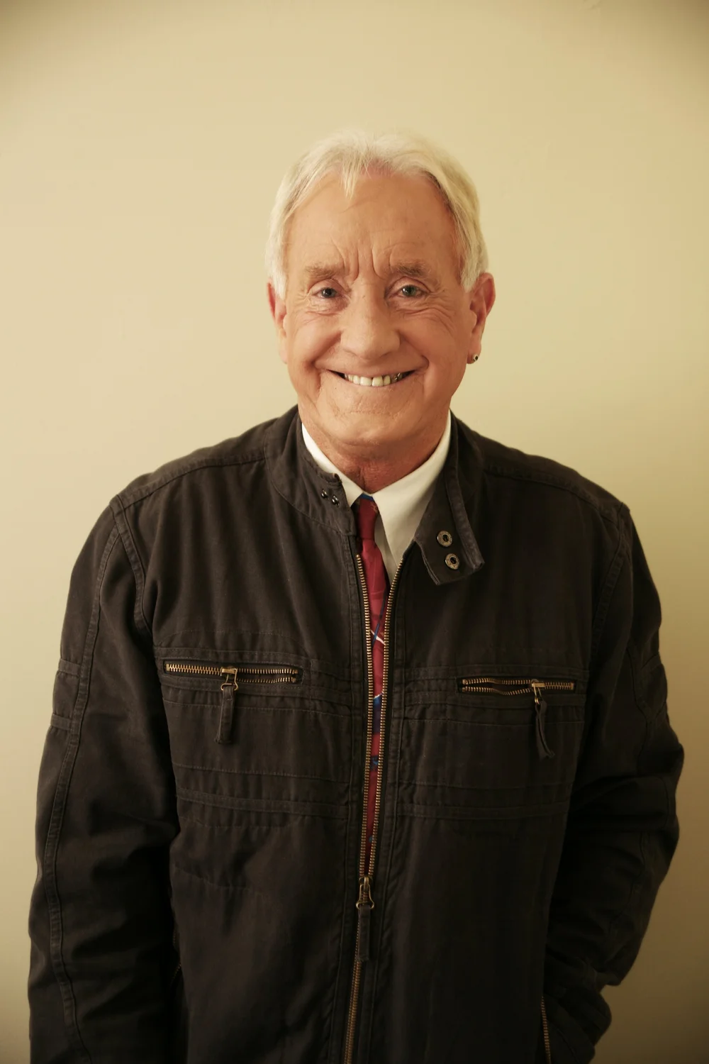 Elderly man smiling in a dark jacket against a light background.
