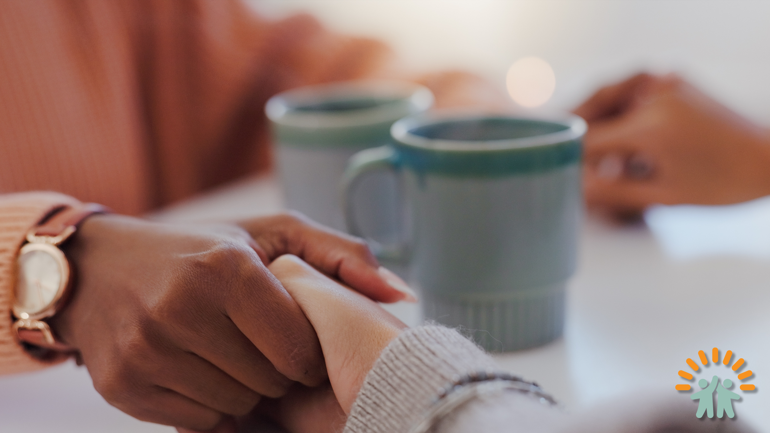 A close-up of two people holding hands on a white surface, with two ceramic mugs in the background, out of focus.