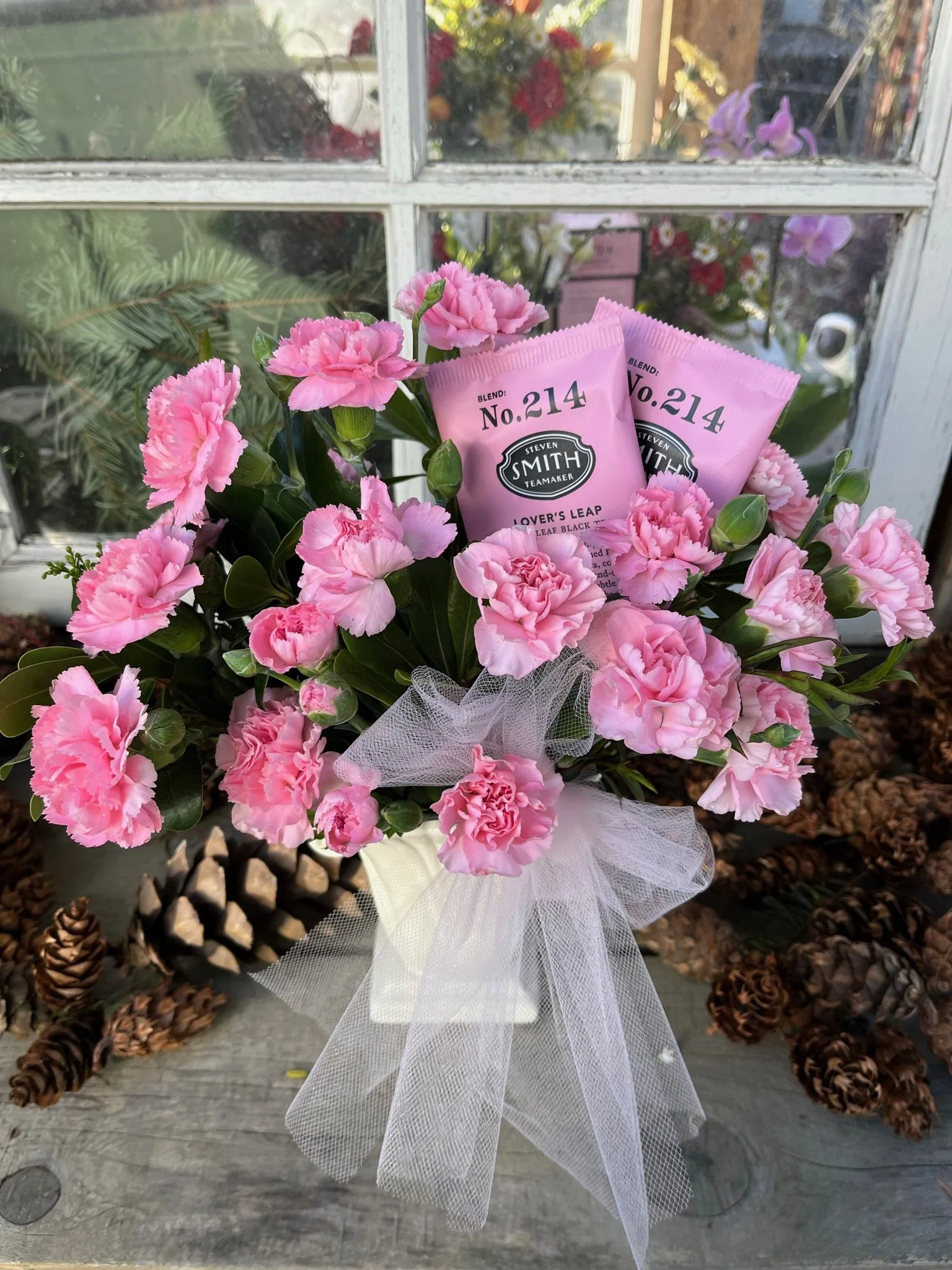 Pink carnation flowers in a white vase with a sheer white bow, two packets of Steven Smith Tea blend No. 214, Lover's Leap, placed among the flowers, wooden surface, pinecones in the background, outside window with reflections of other flowers.