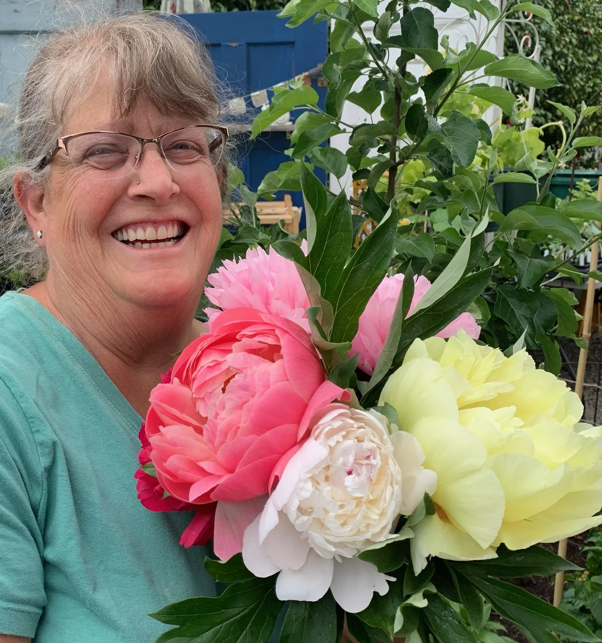 A smiling woman with glasses holding a bouquet of pink, white, and yellow peonies in a garden.