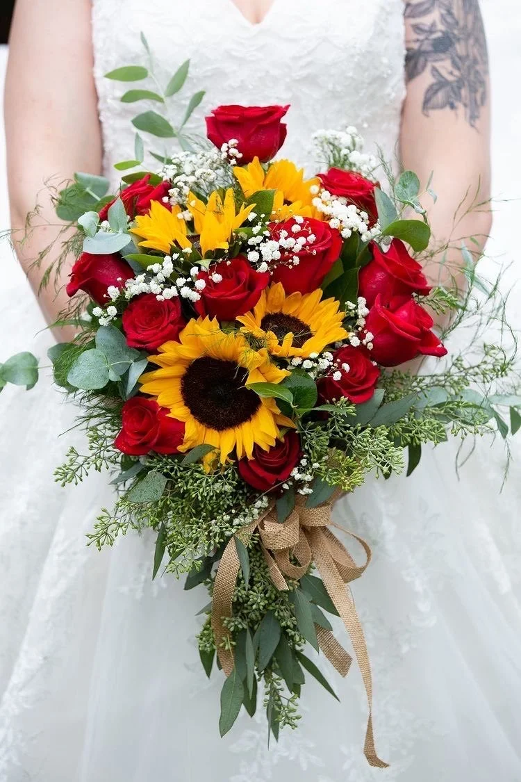 A woman in a white dress holding a bouquet of red roses, sunflowers, baby’s breath, and greenery, tied with a brown ribbon.