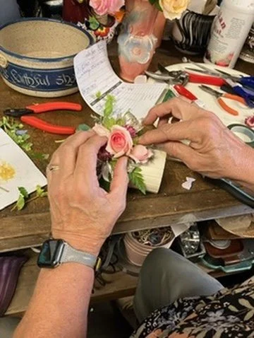 Person arranging pink roses and greenery on a craft project at a cluttered work table.