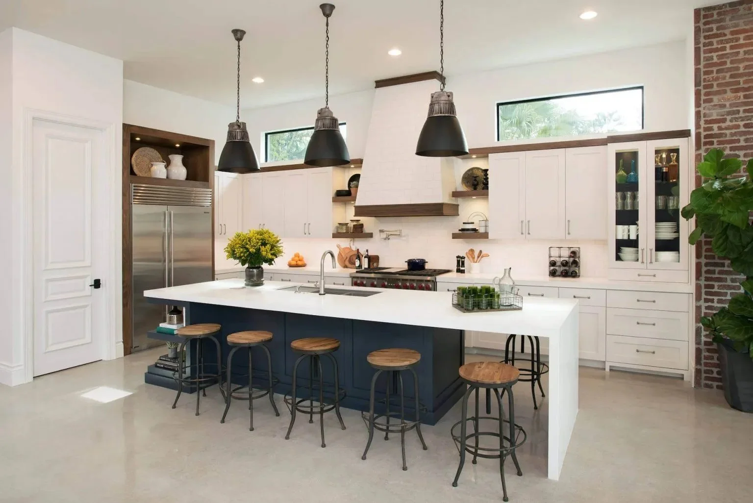 Modern kitchen with white cabinets, a navy blue island with wooden stools, exposed brick wall, and black pendant lighting.