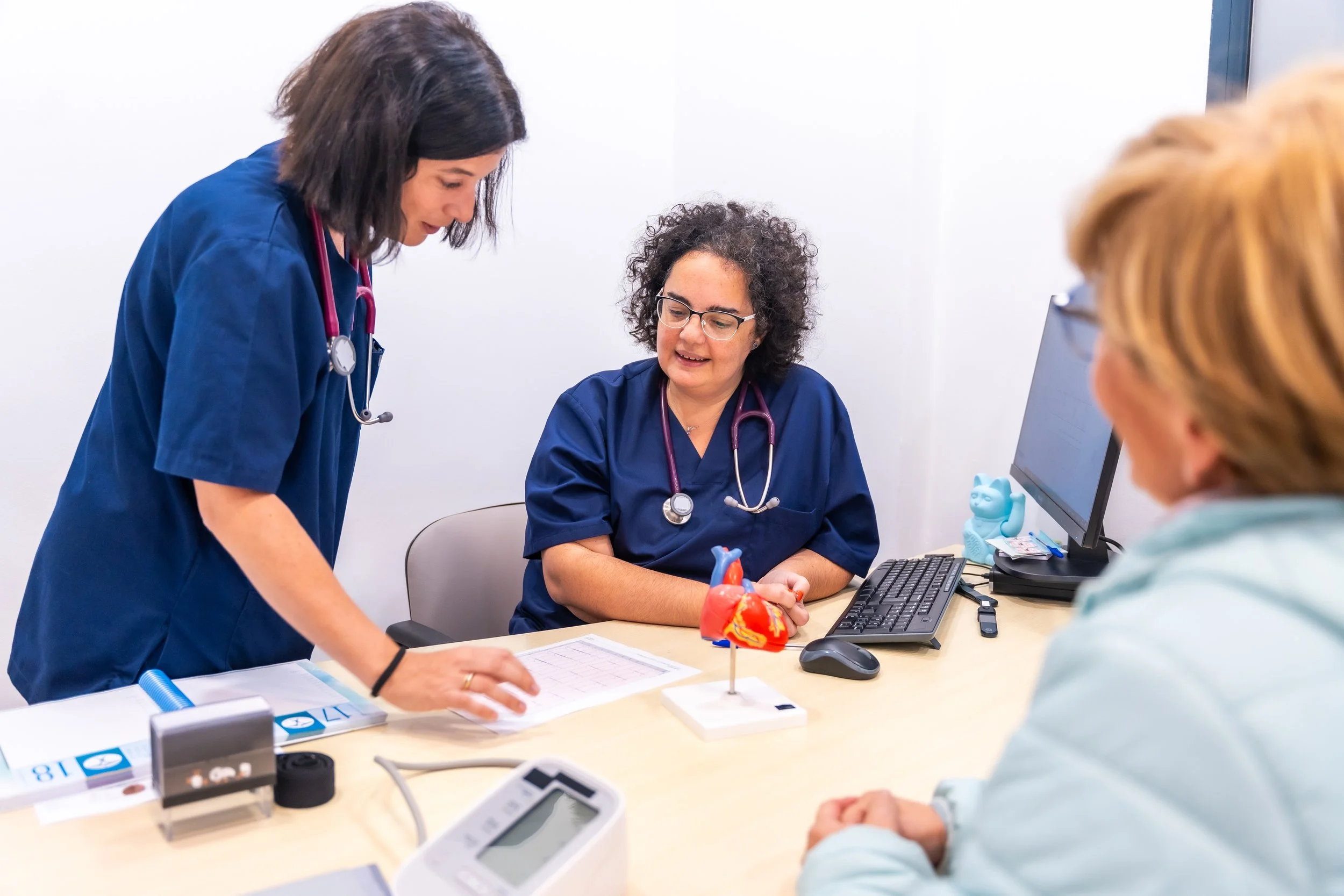 Two female nurses in blue scrubs discuss with a female patient in a medical office, with a model of a heart on the desk.
