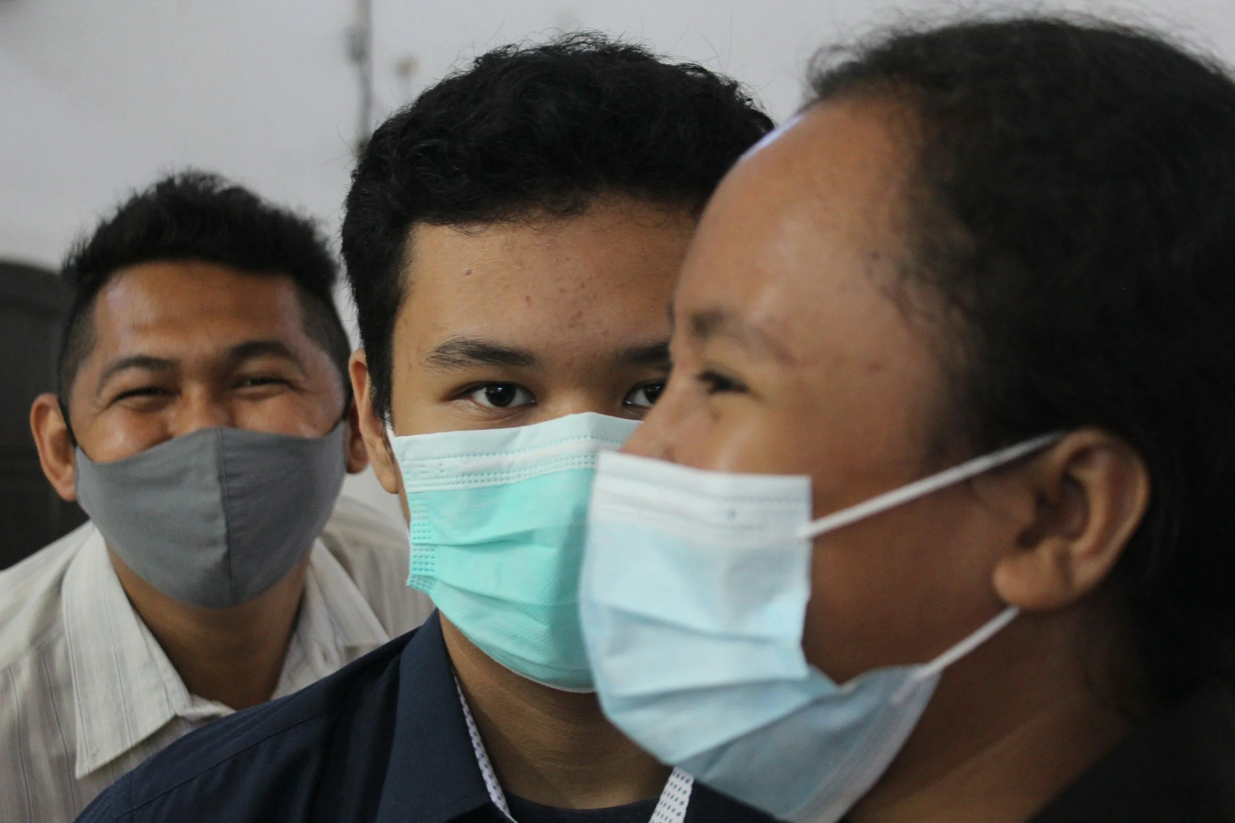 Three people wearing face masks, standing close together, engaged in conversation. The person in the middle has intense eyes directly at the camera, while the others look at each other with smiling eyes, indicating a friendly interaction.