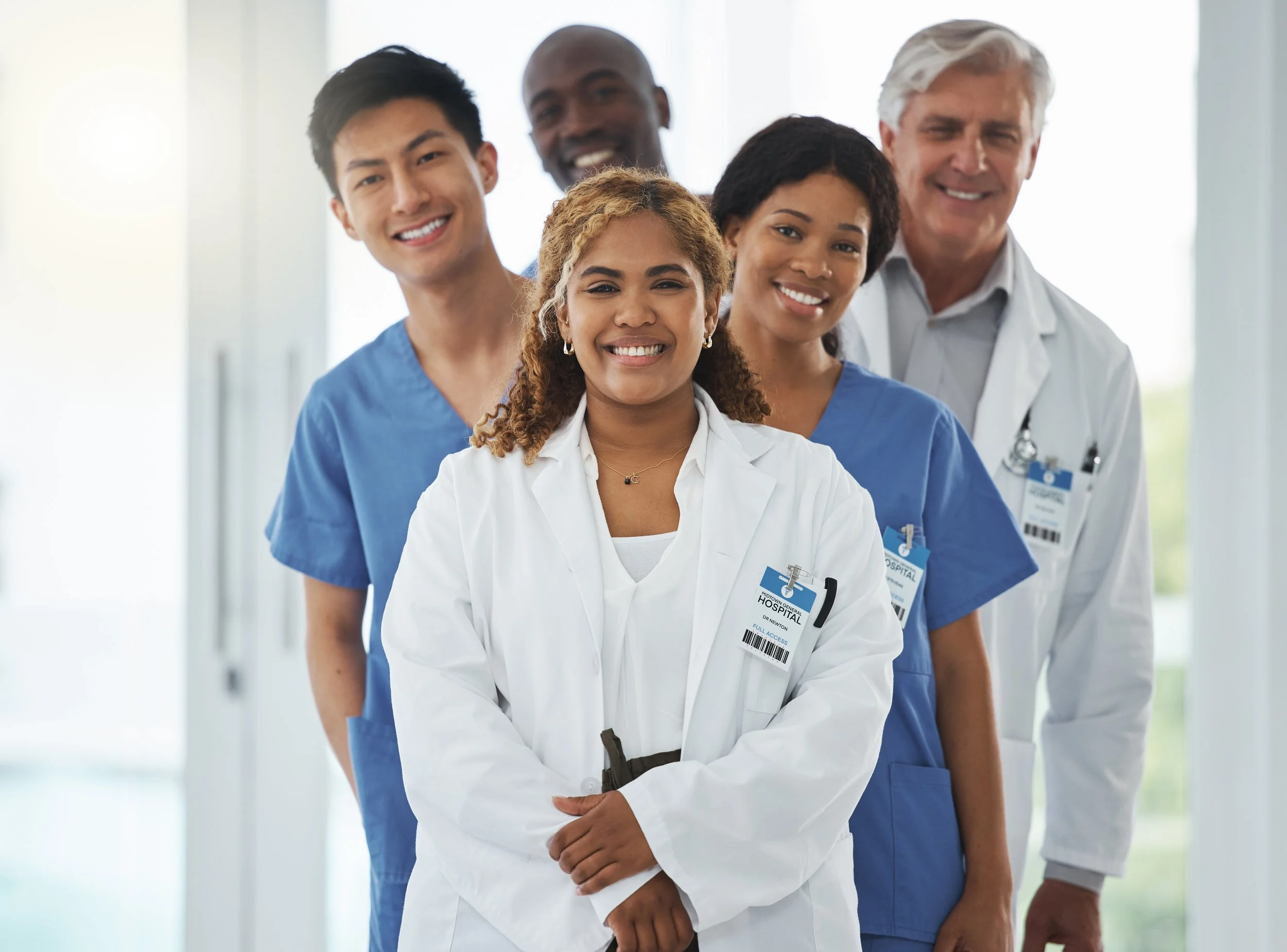 Group of five diverse medical professionals standing together in hospital hallway, smiling, wearing scrubs and white coats with ID badges.