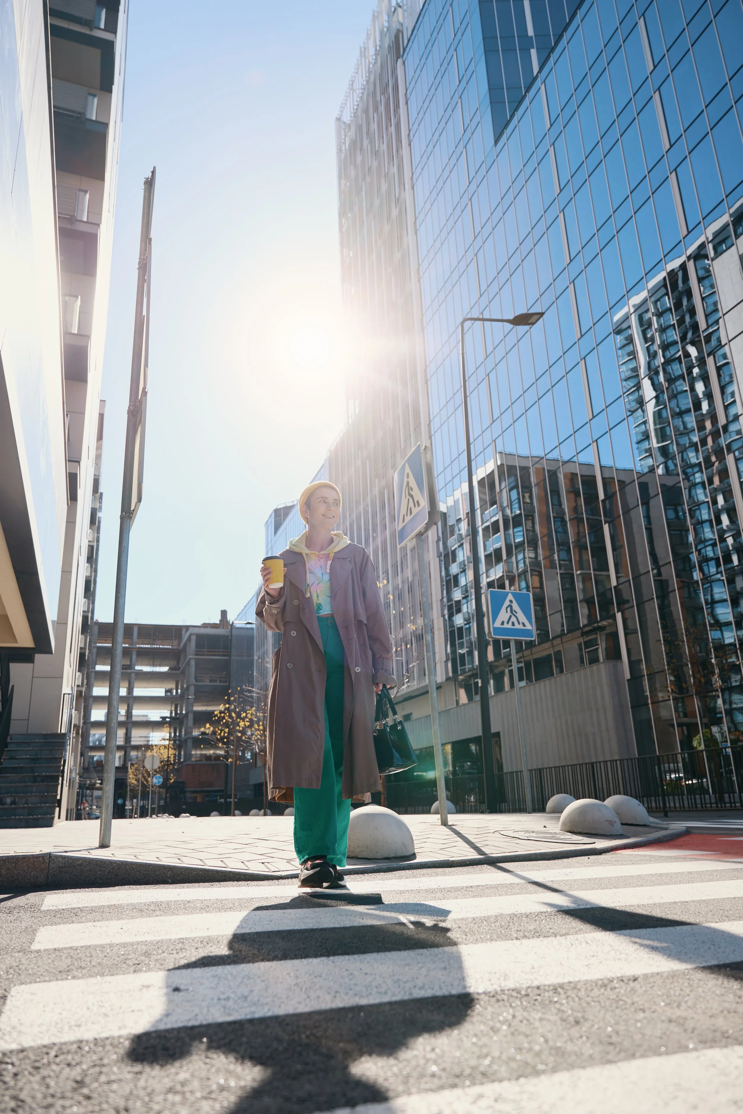 A woman walking across a city crosswalk holding a coffee cup and a handbag with modern glass skyscrapers reflecting the sky and sunlight in the background.