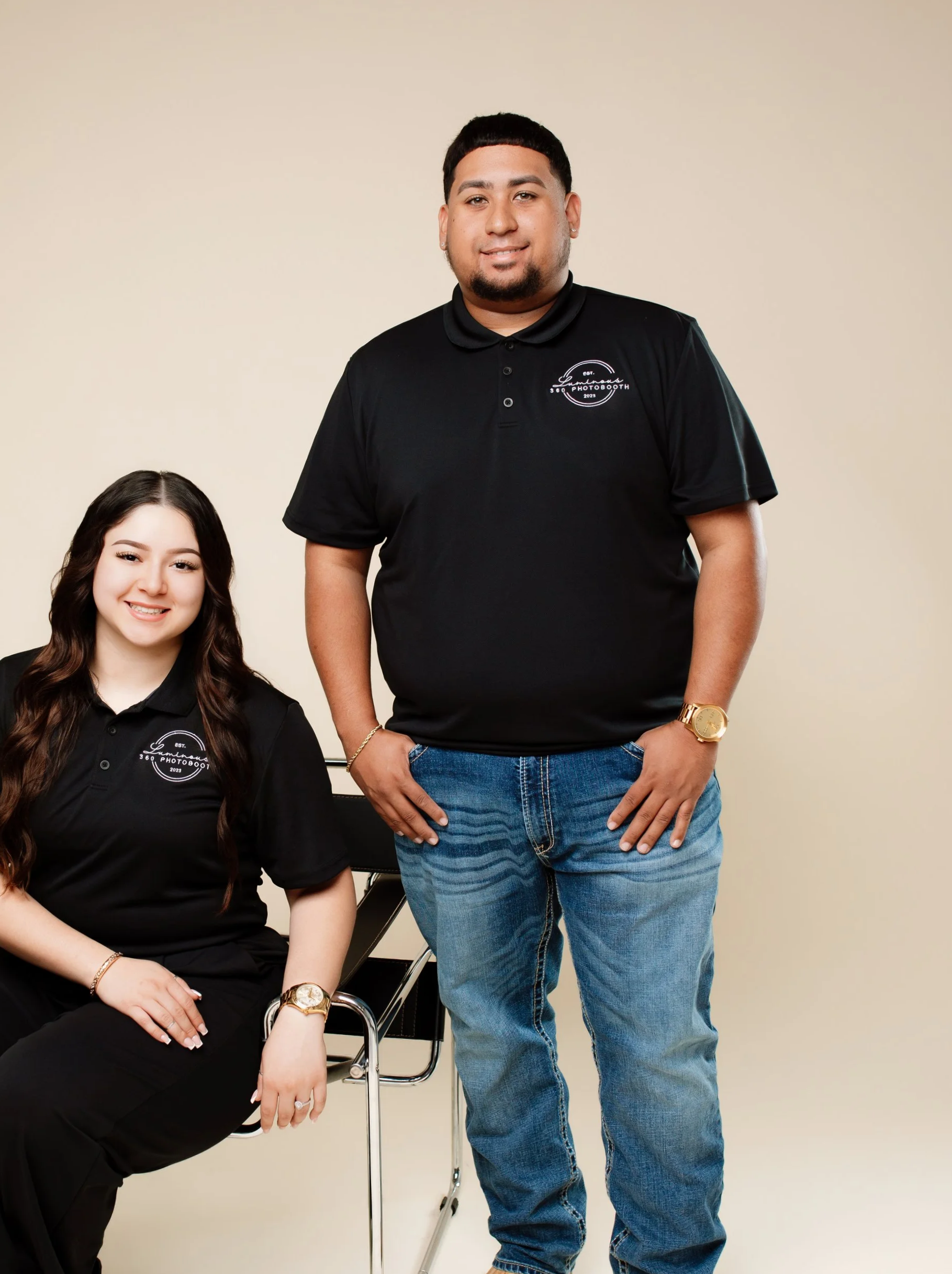 A young woman sitting on a chair and a young man standing next to her, both wearing black polo shirts with a logo and text that reads 'Luminous 360 Photobooth EST 2023'.