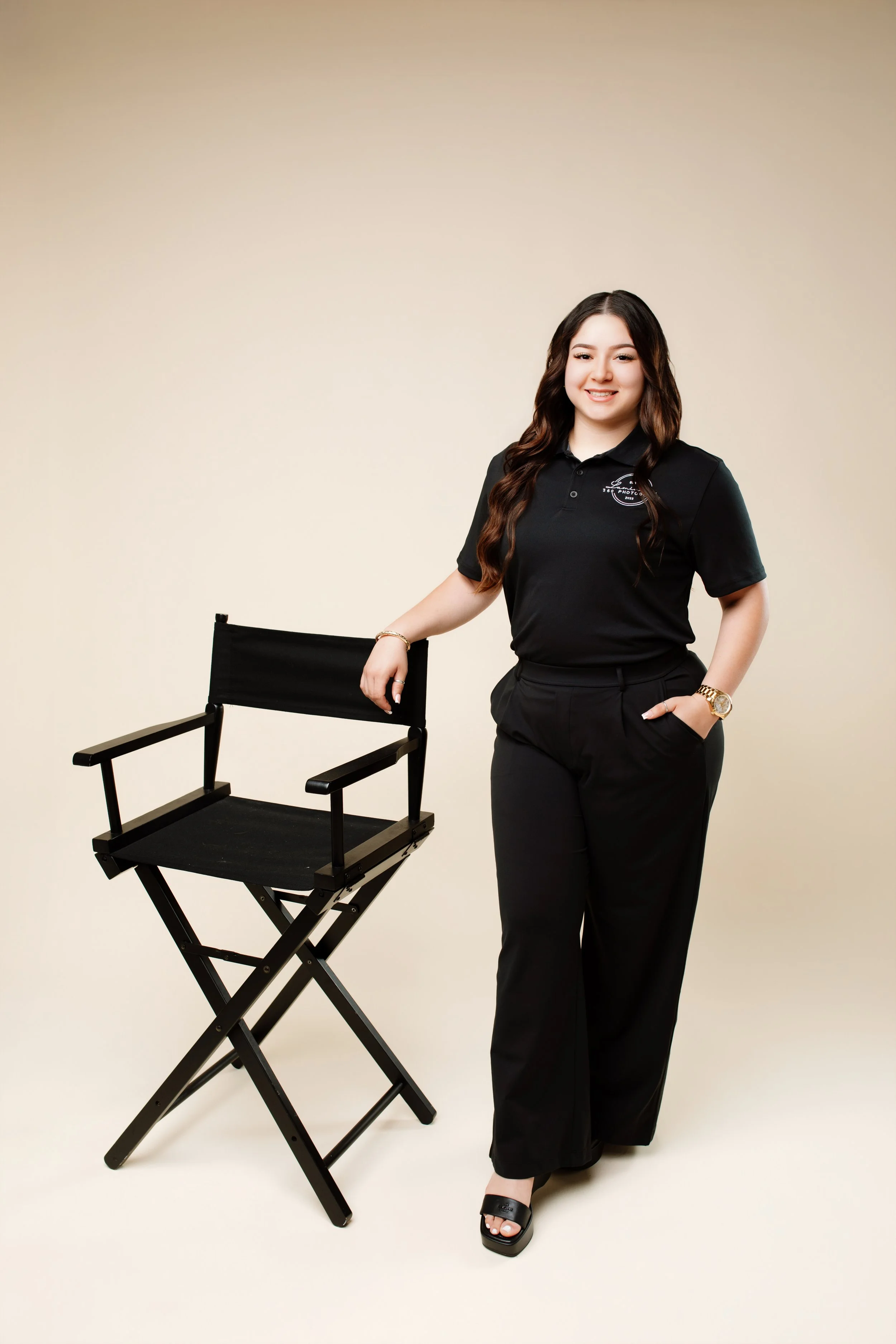A young woman with long wavy brown hair dressed in black pants and a black polo shirt standing next to a black director's chair, smiling at the camera, in a studio setting with a plain beige background.
