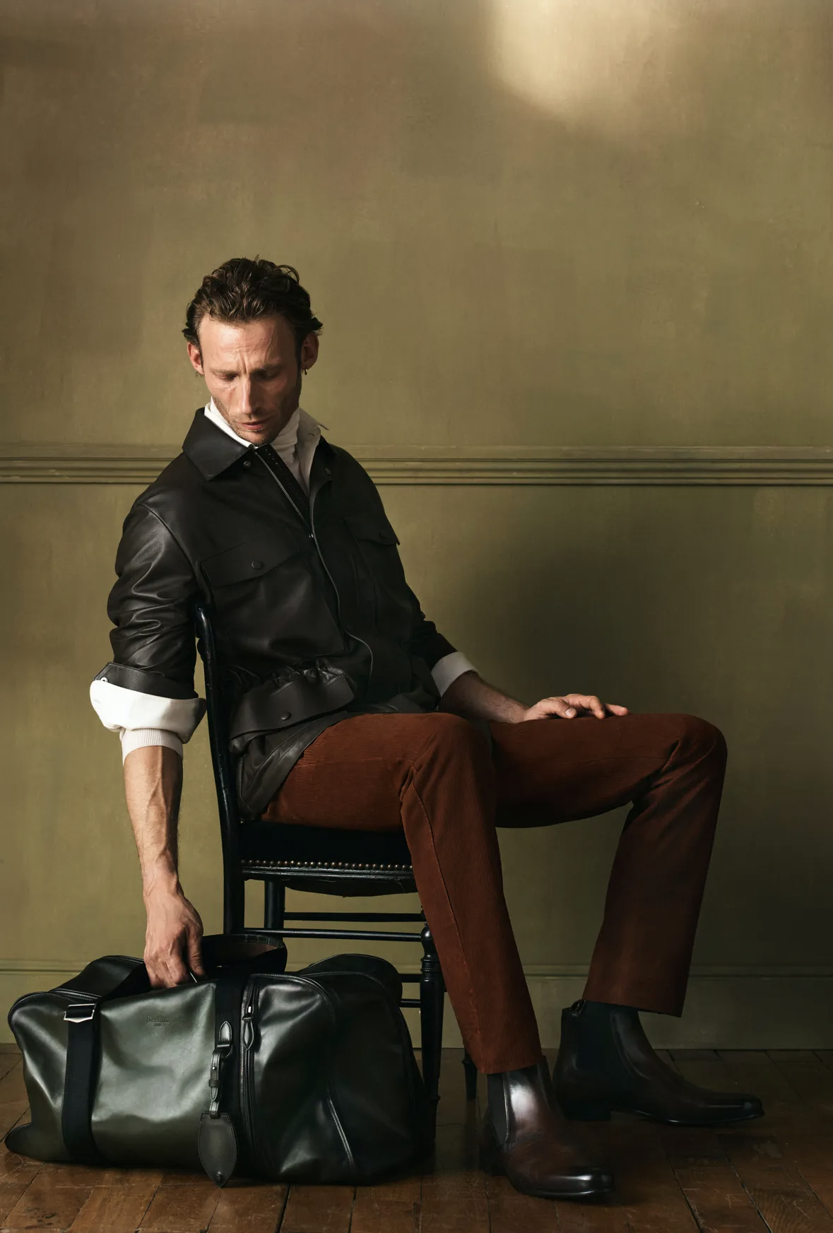 Seated man in a black leather jacket and rust trousers with a leather travel bag