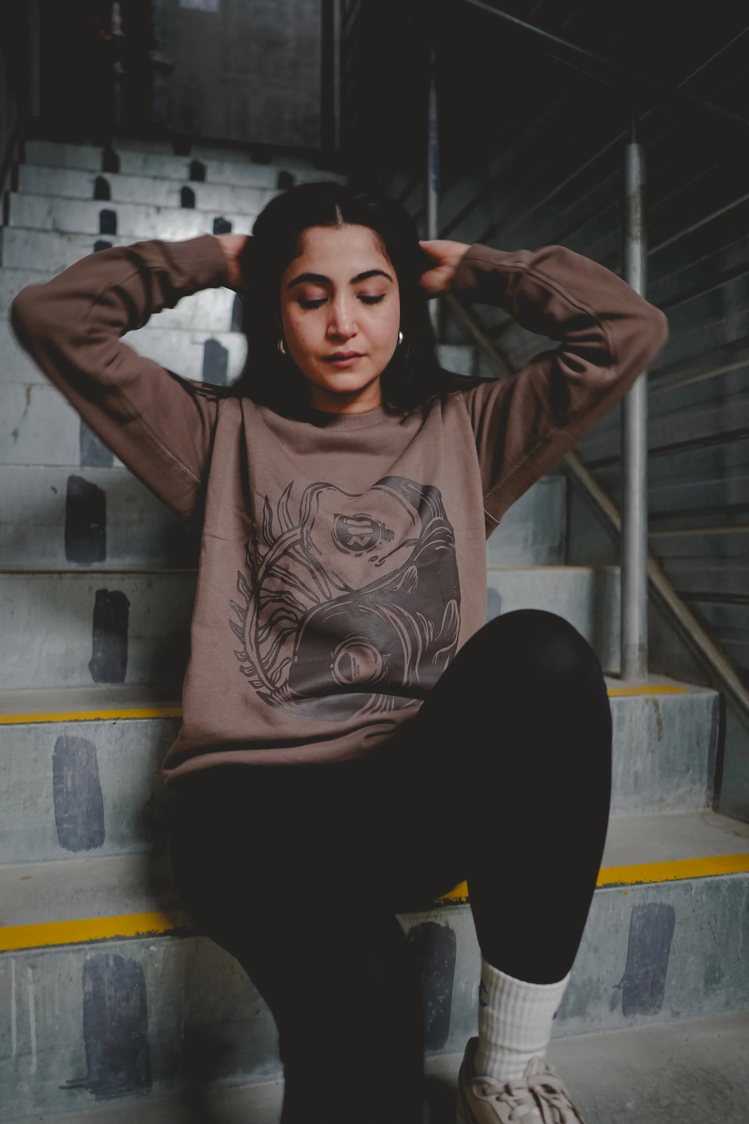 A young woman with dark hair sitting on concrete stairs, wearing a brown sweatshirt with a graphic design and black pants, with her hands behind her head and eyes closed.