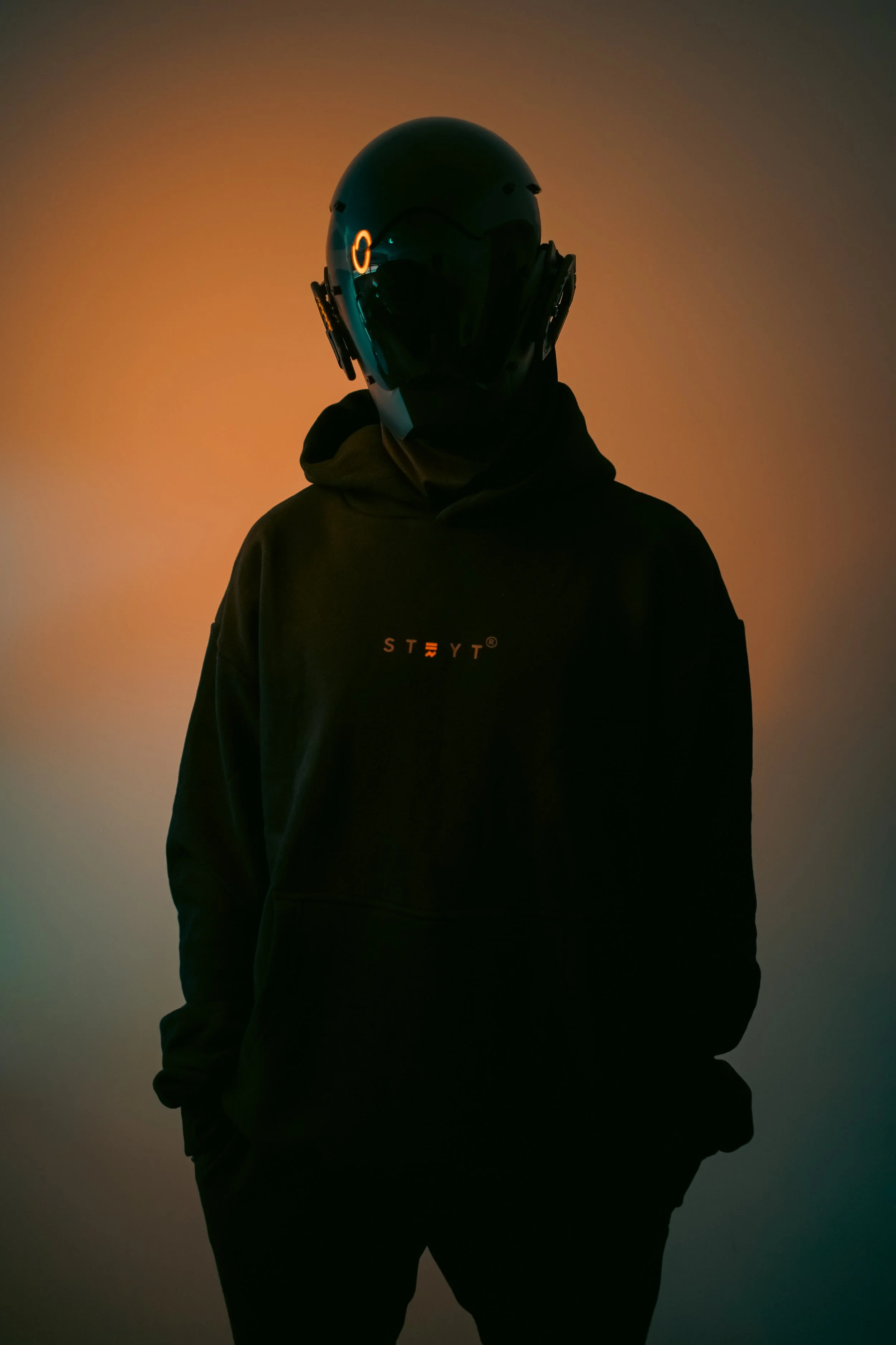 A person wearing a black hoodie with the logo 'STRYT' on the front and a futuristic helmet, standing against a multicolored gradient background.