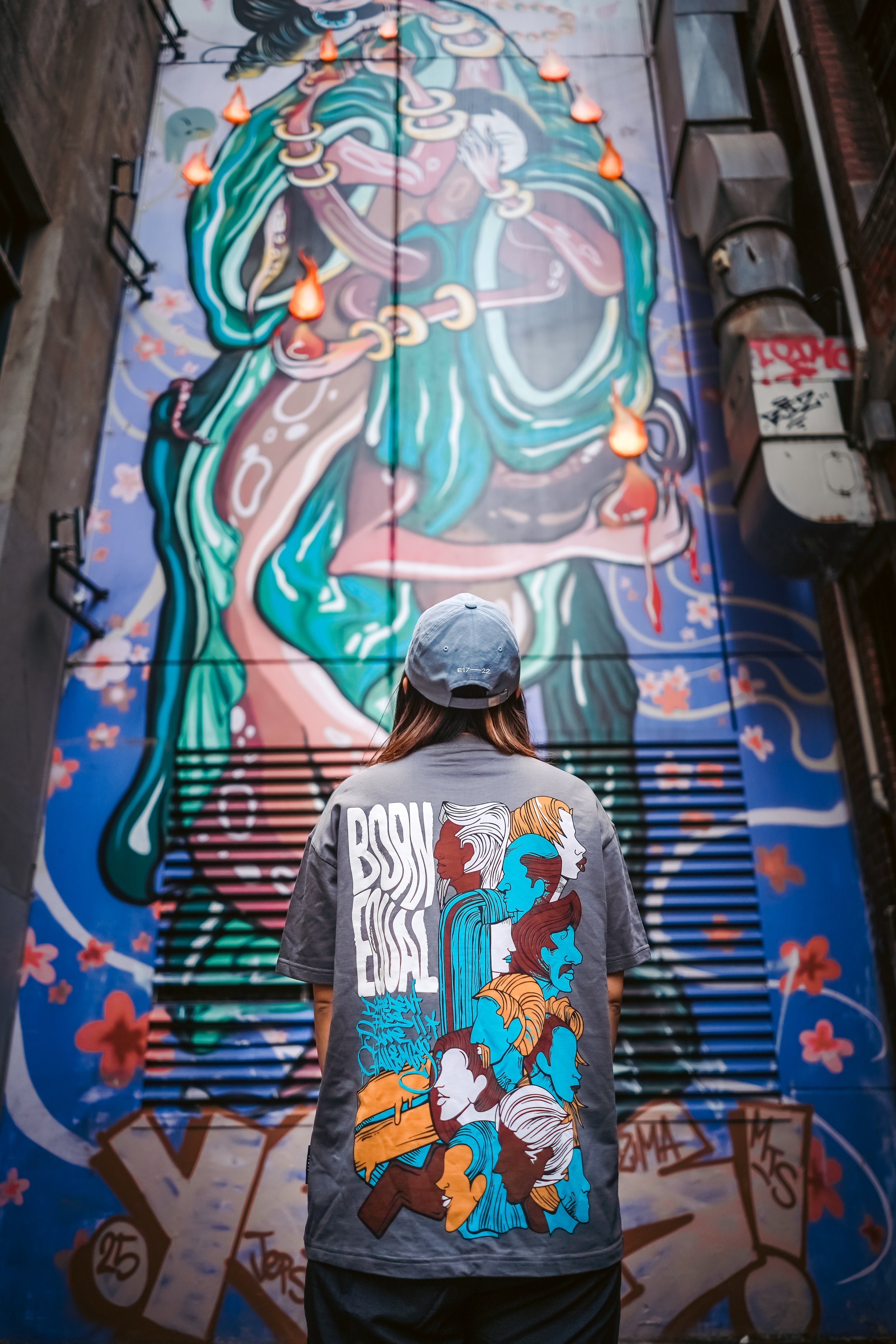 Person wearing a gray T-shirt with illustrated faces of women in various colors walking in front of a large, colorful mural of a woman with long flowing hair and multiple orange flames hanging from her head.