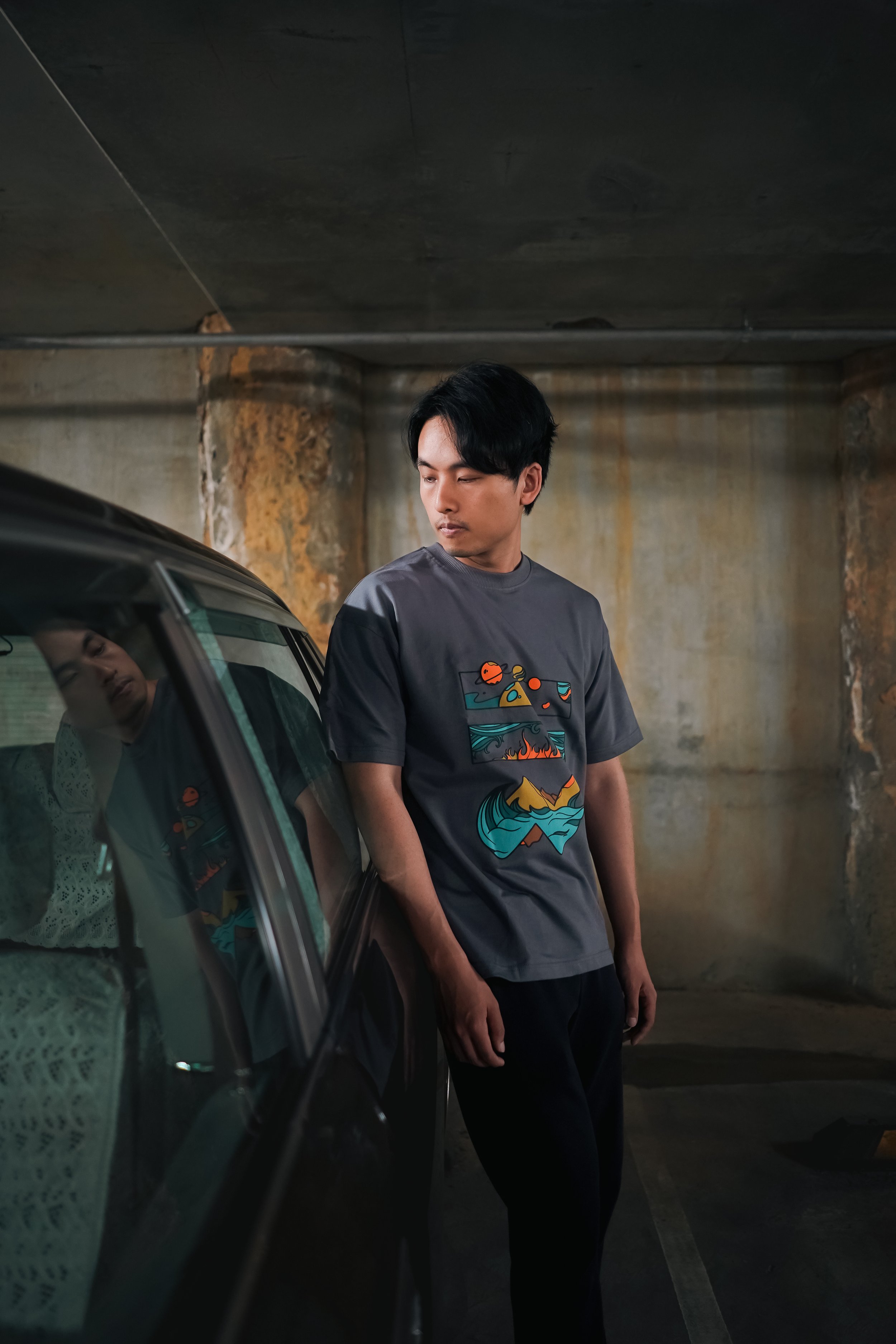 Young man with black hair wearing a gray graphic T-shirt, standing in an underground parking garage, leaning against a black car, with concrete and rusty walls in the background.