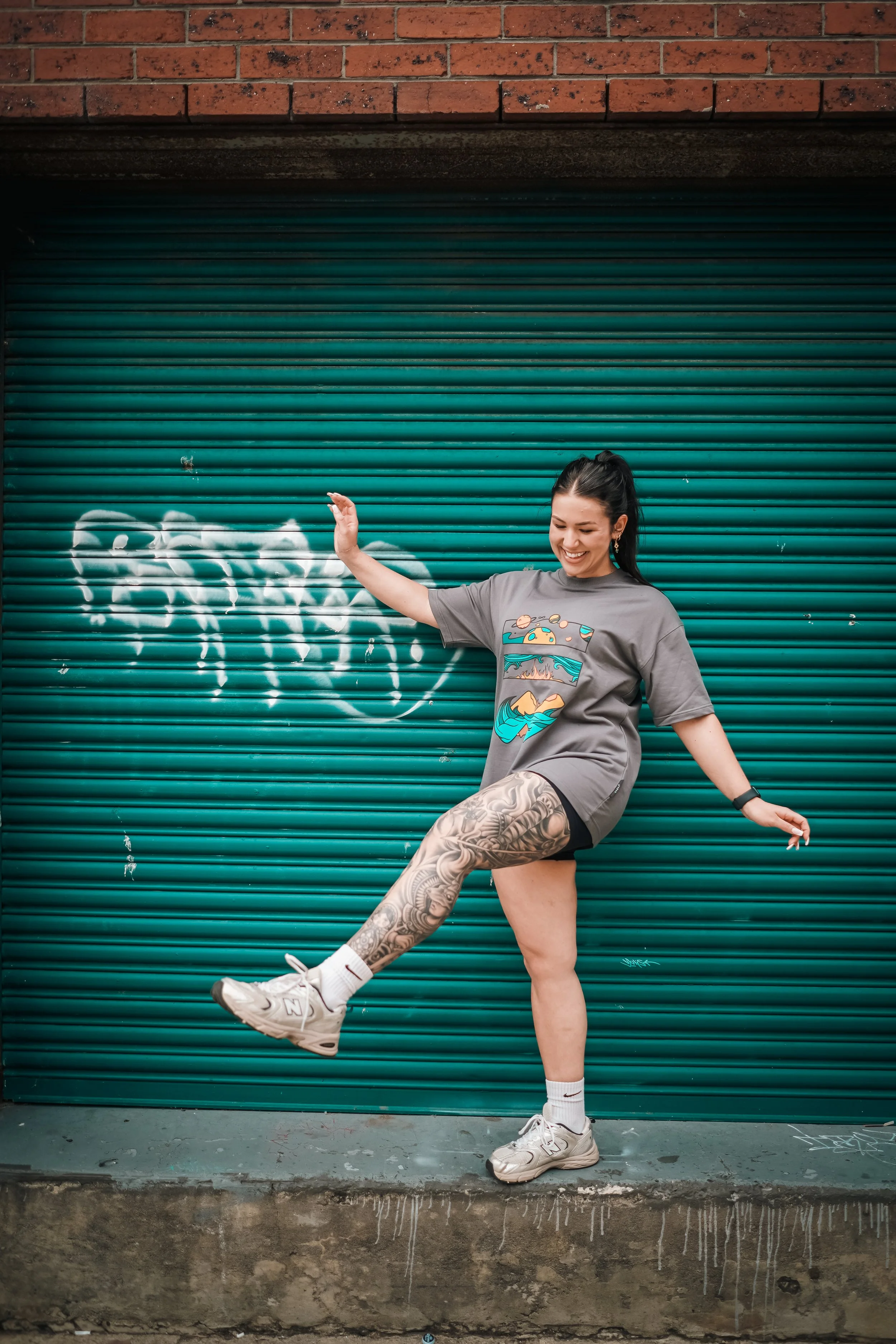 A woman in a gray t-shirt with a colorful graphic, black shorts, and white sneakers, standing against a teal graffiti-covered metal door, smiling and pretending to kick.