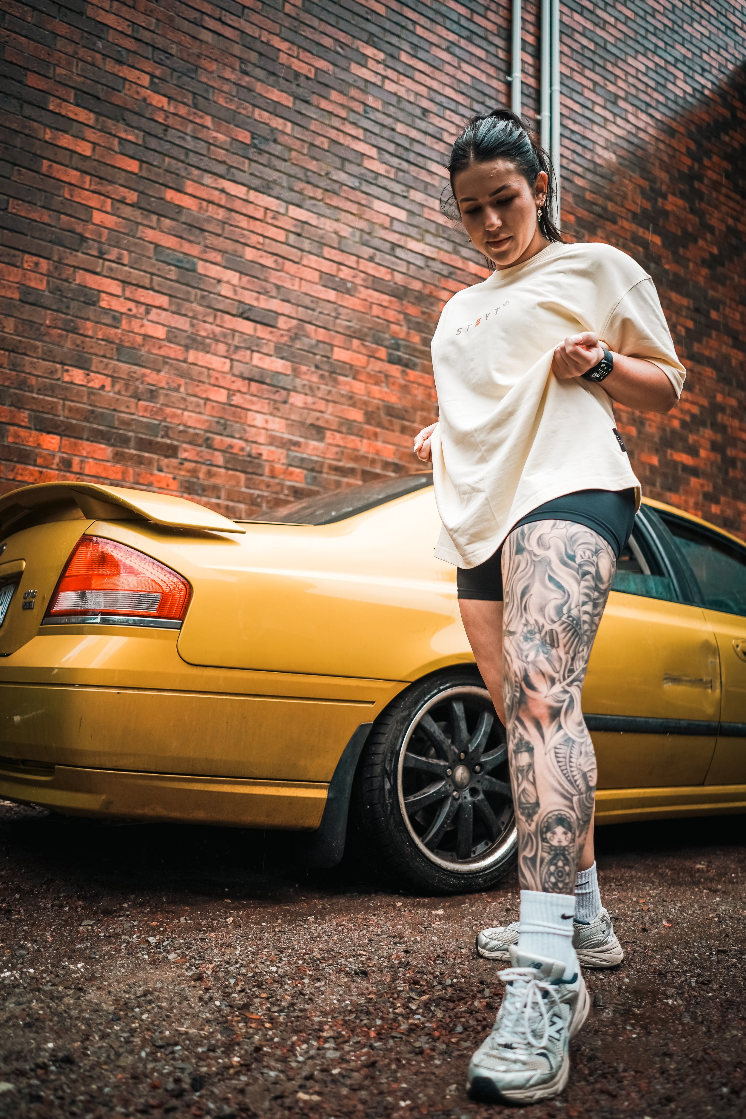 A woman with tattoos on her leg stands in front of a yellow car with a brick wall background, adjusting her oversized beige t-shirt.