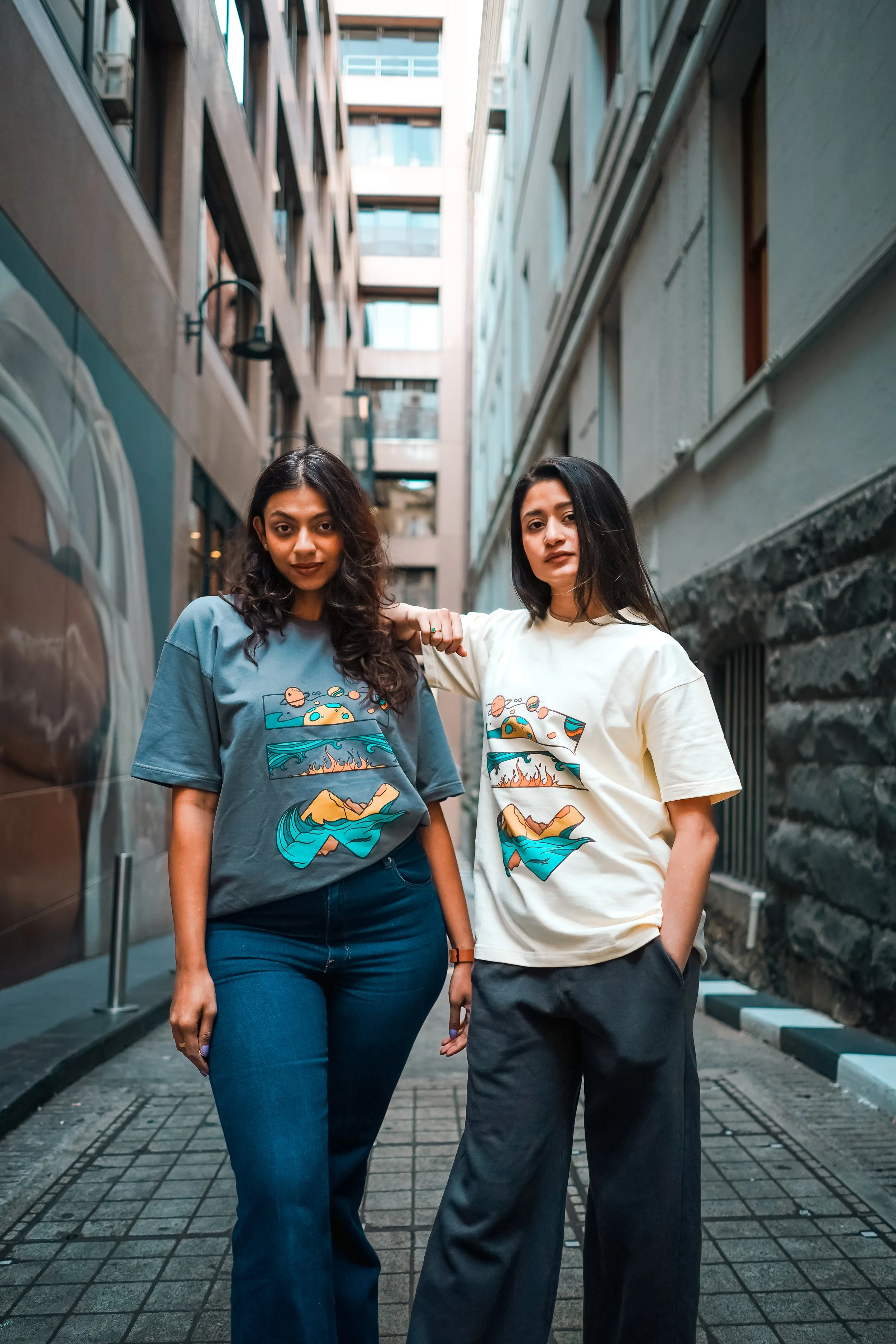 Two women standing in a narrow city alleyway, wearing graphic T-shirts with landscape designs, posing with relaxed expressions.