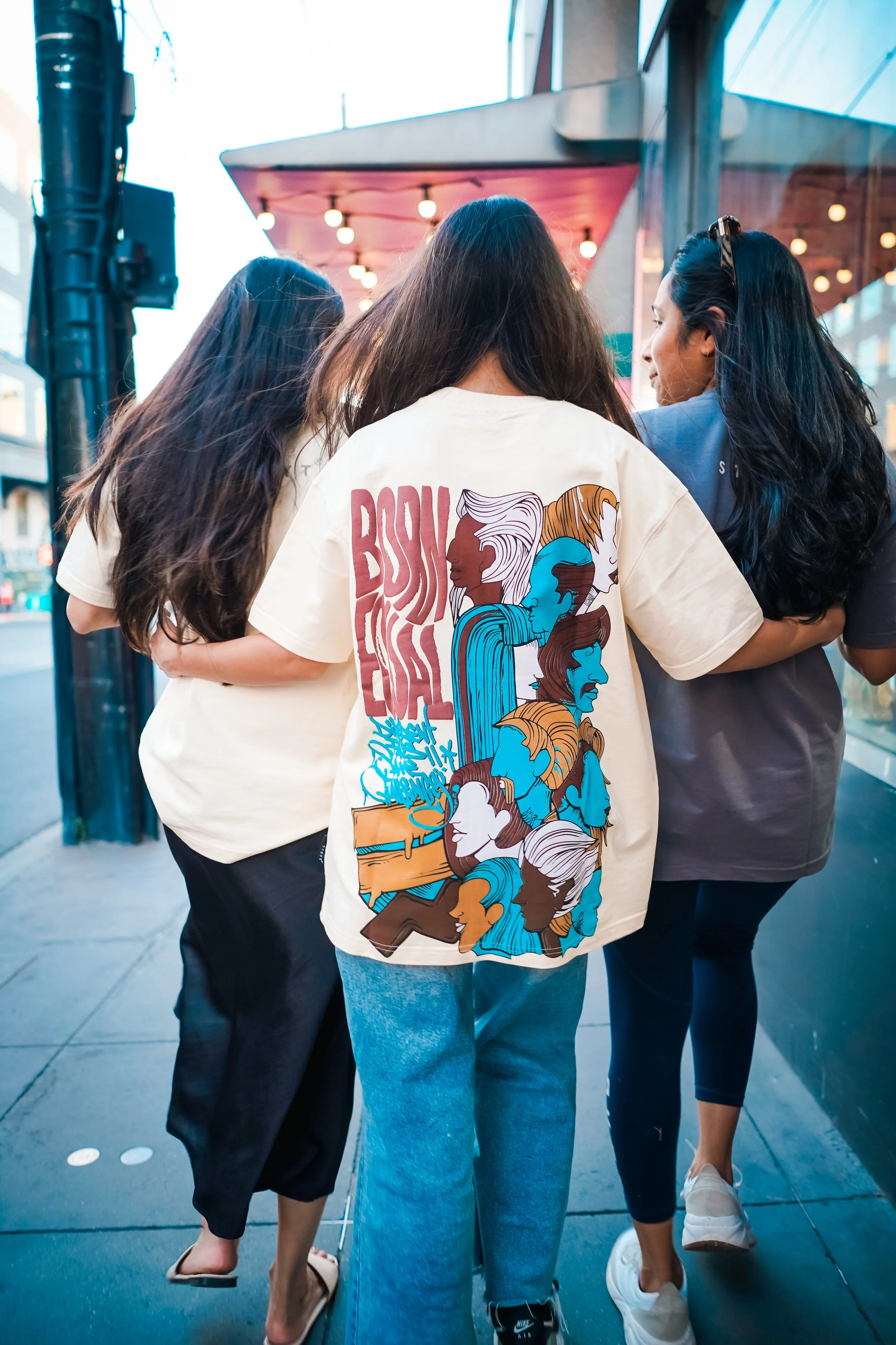 A group of four women walking together on a city sidewalk at dusk, with colorful streetlights and buildings around them. They are dressed casually, and one woman in the center is wearing a cream-colored T-shirt with colorful illustrations of diverse 