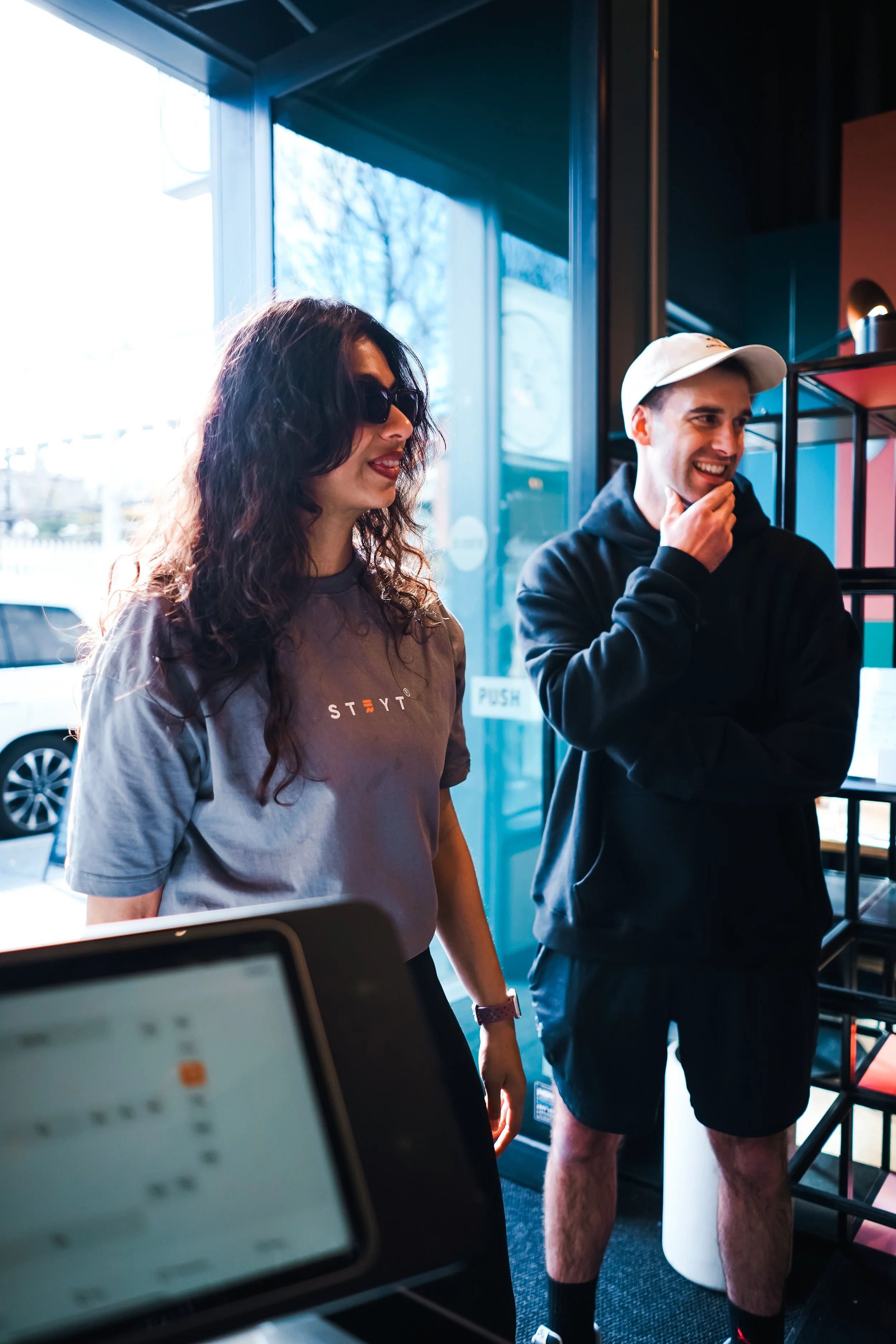 Two young people standing inside a modern store near the window, smiling and talking, with a laptop in the foreground and shelves behind them.