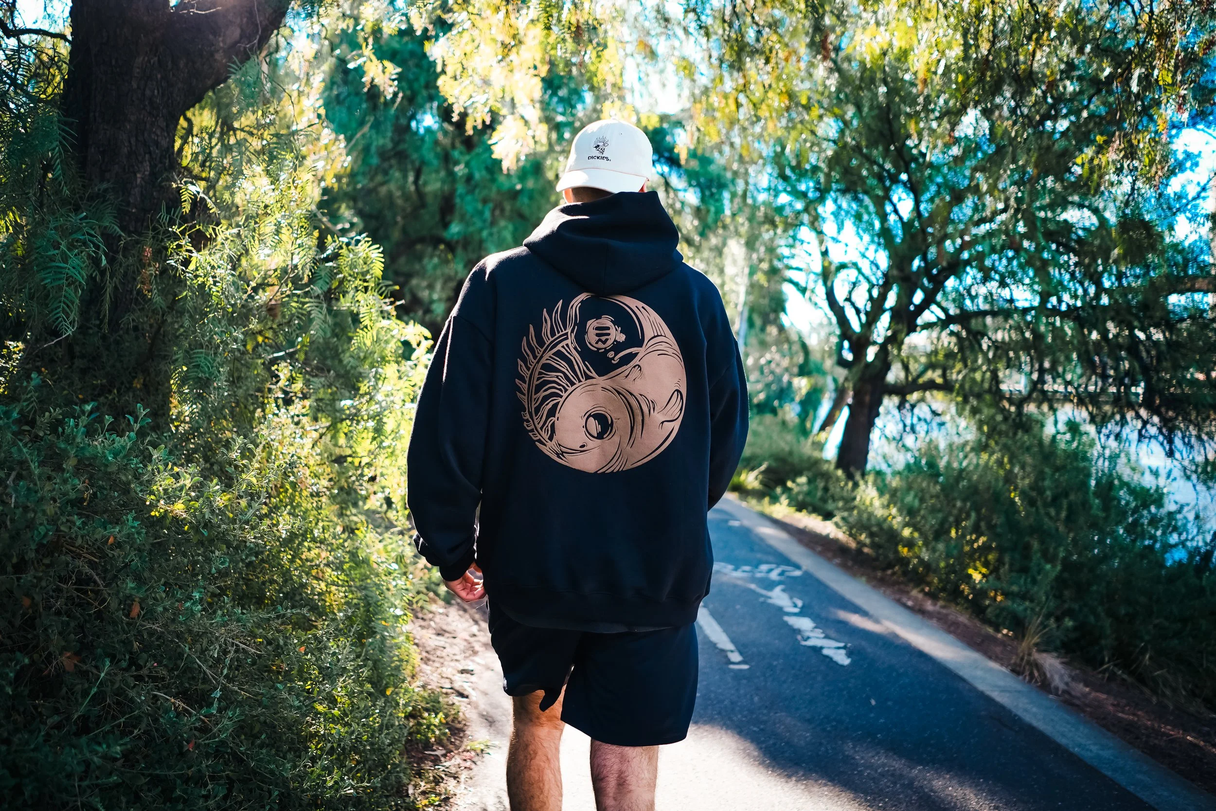 A person walking on a trail surrounded by trees, wearing a black hoodie with a gold yin-yang design on the back with sunlight filtering through the foliage.