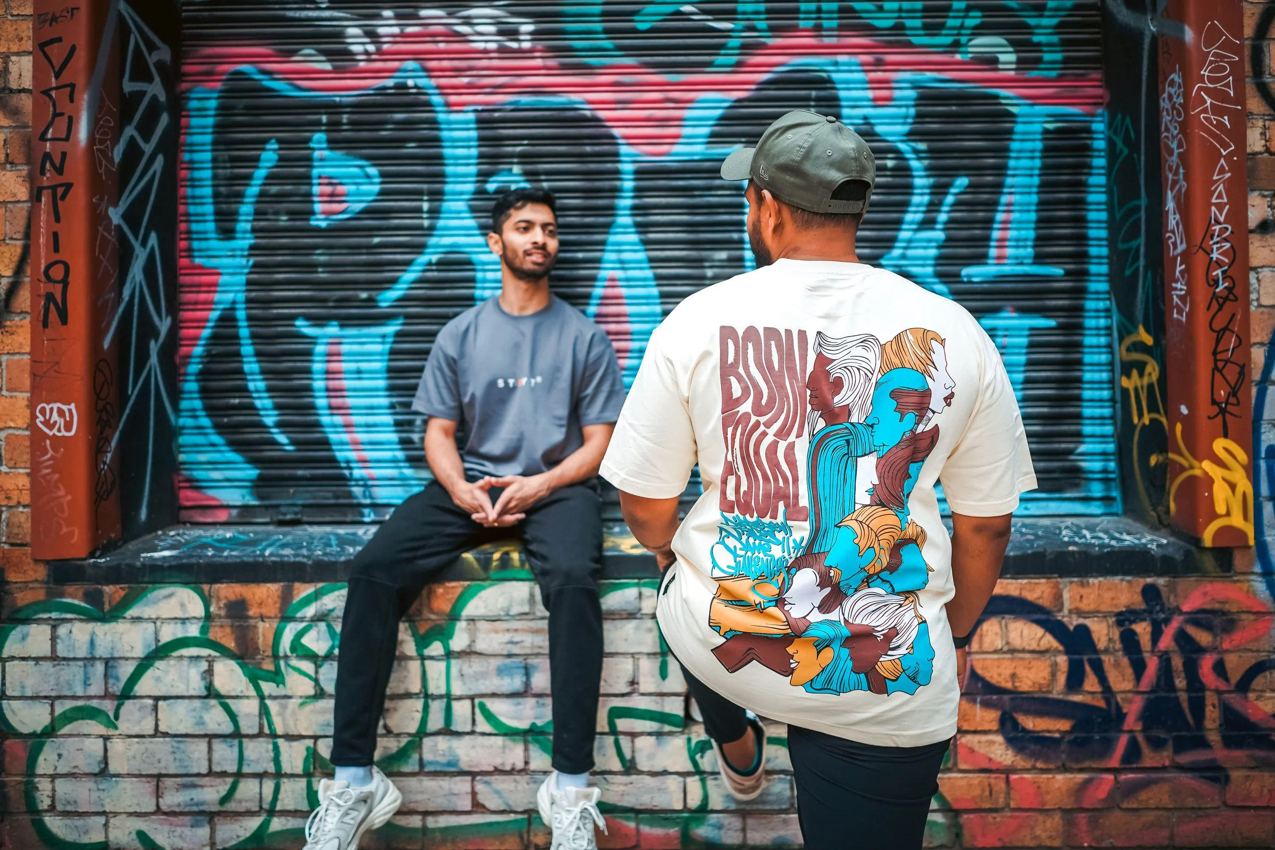 Two young men in front of a graffiti-covered wall, one sitting on a ledge and the other standing, engaged in conversation.