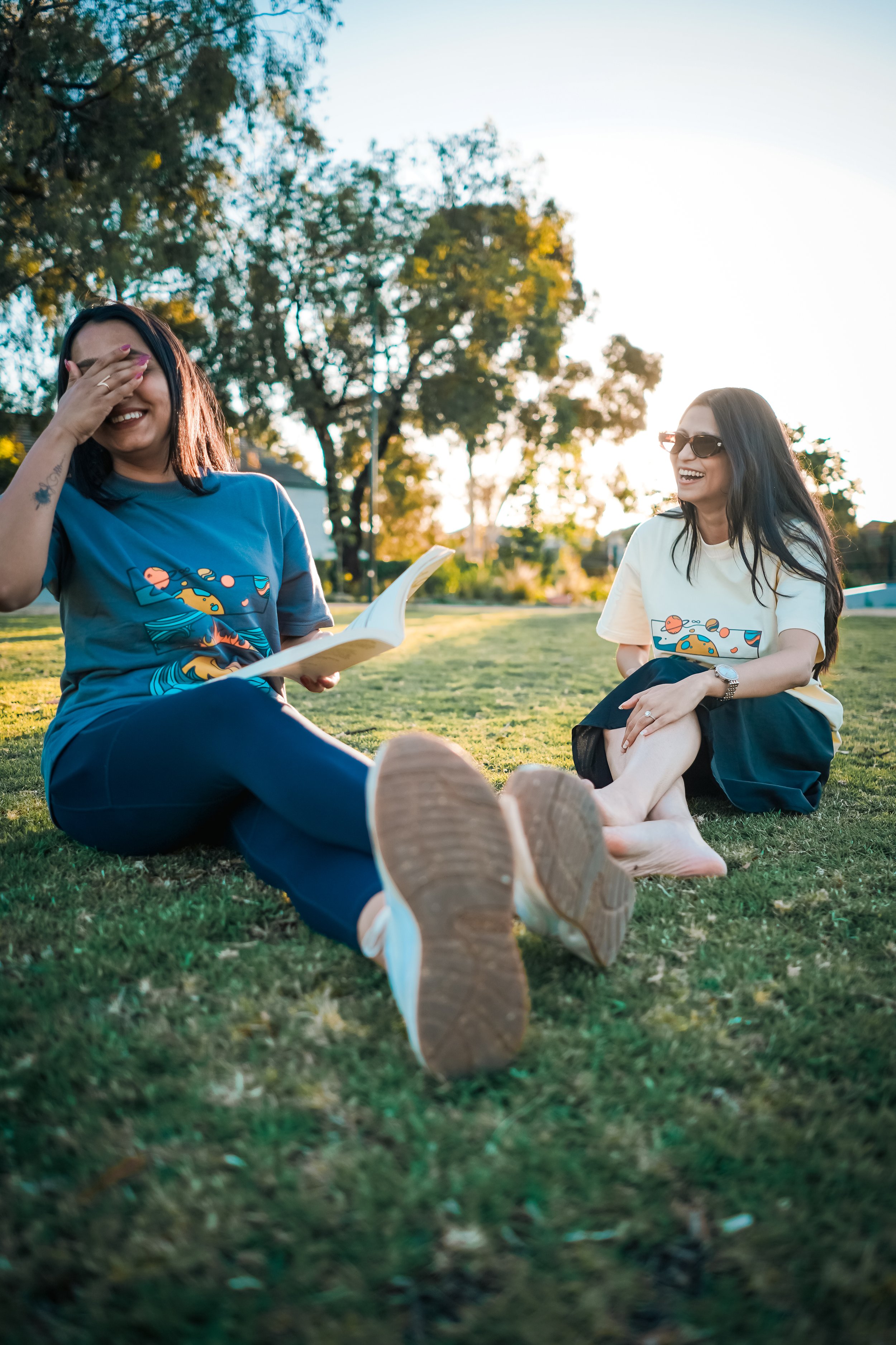 Two women sitting on grass in a park, one laughing and covering her face, the other smiling, both wearing casual clothes and sunglasses, enjoying a sunny day.
