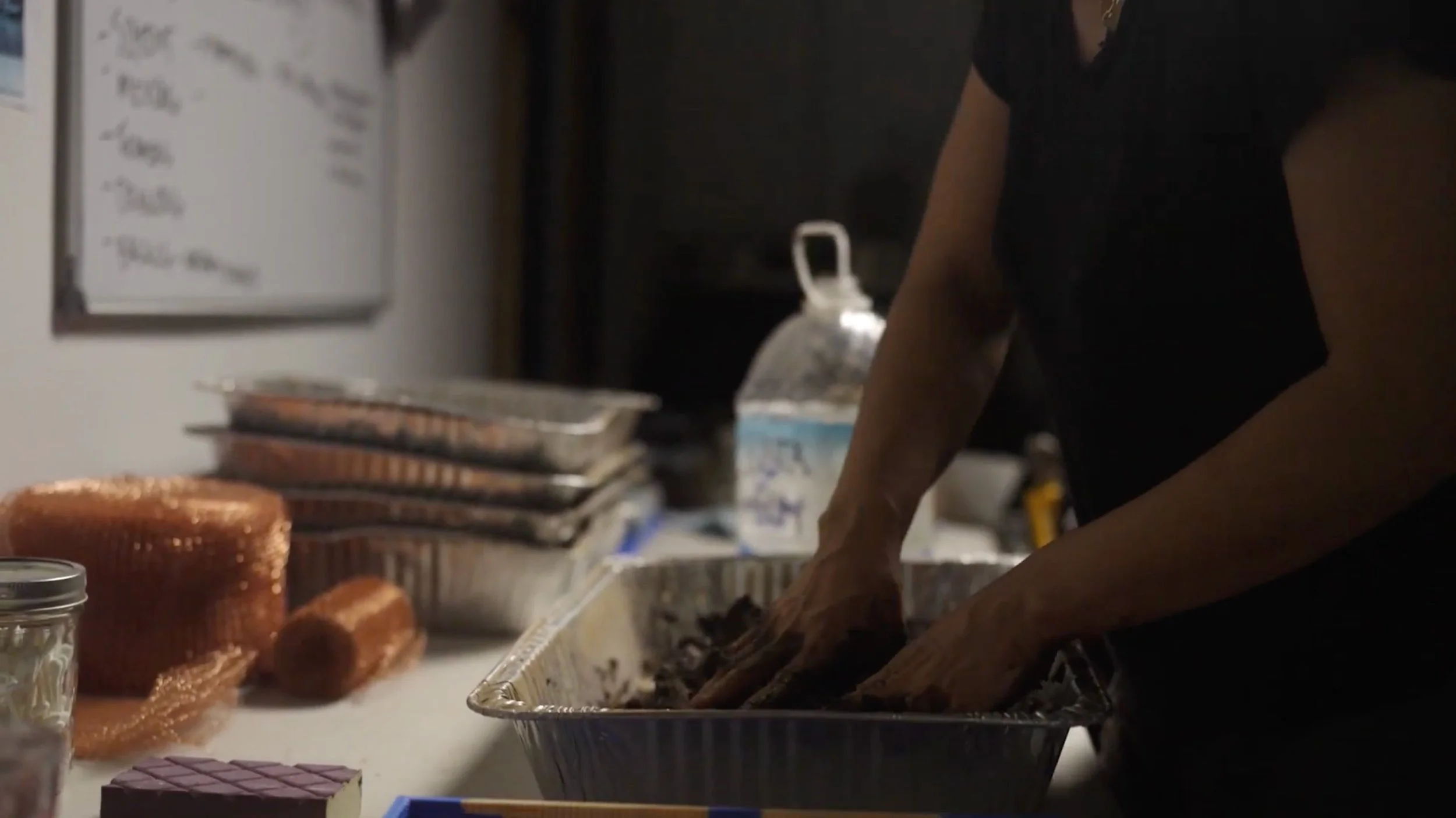 A person preparing food at a kitchen counter with a foil tray, food containers, and baked goods nearby.