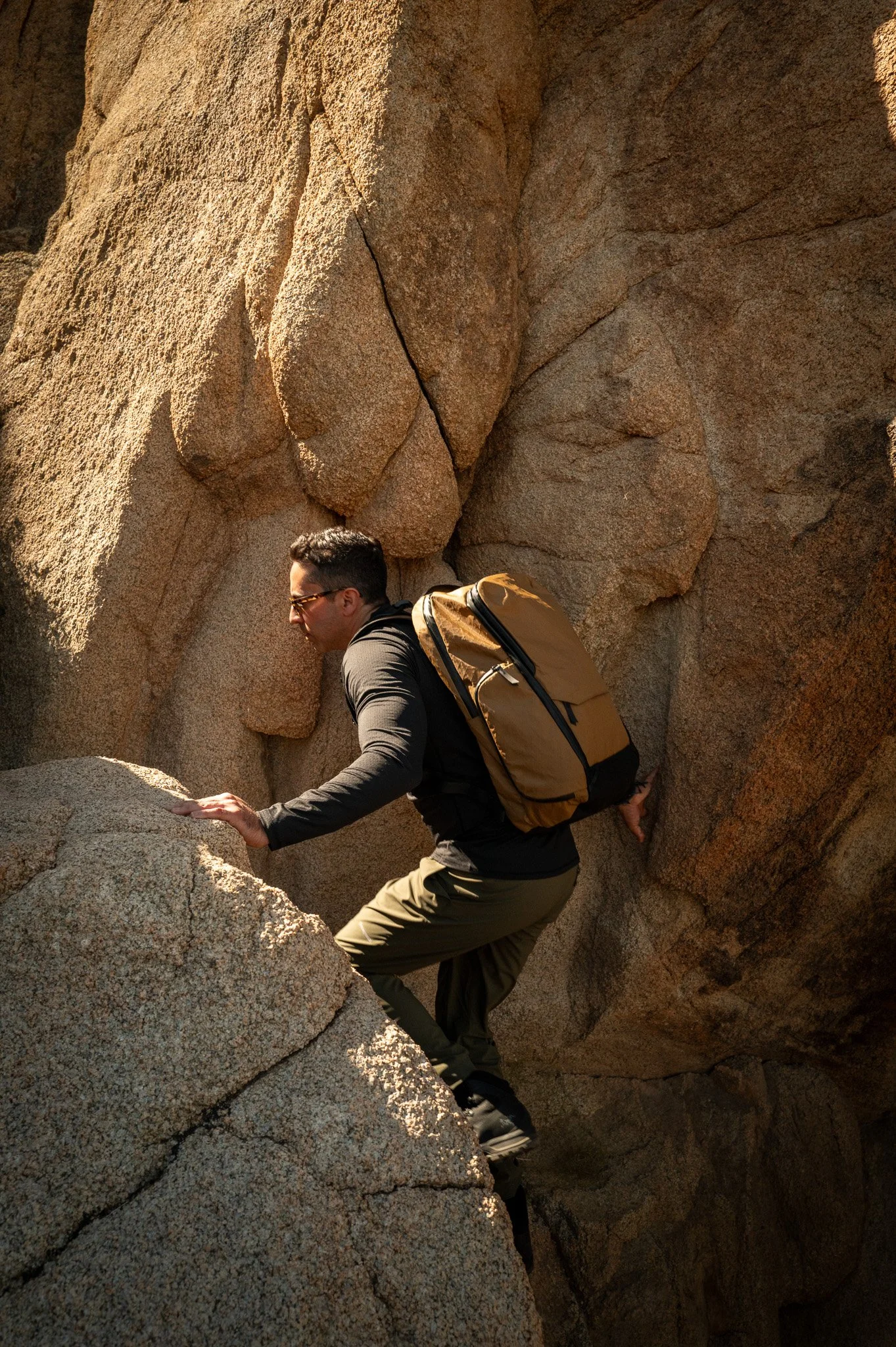 A man wearing glasses, a black long-sleeve shirt, khaki pants, and a yellow backpack is rock climbing on a large granite formation.