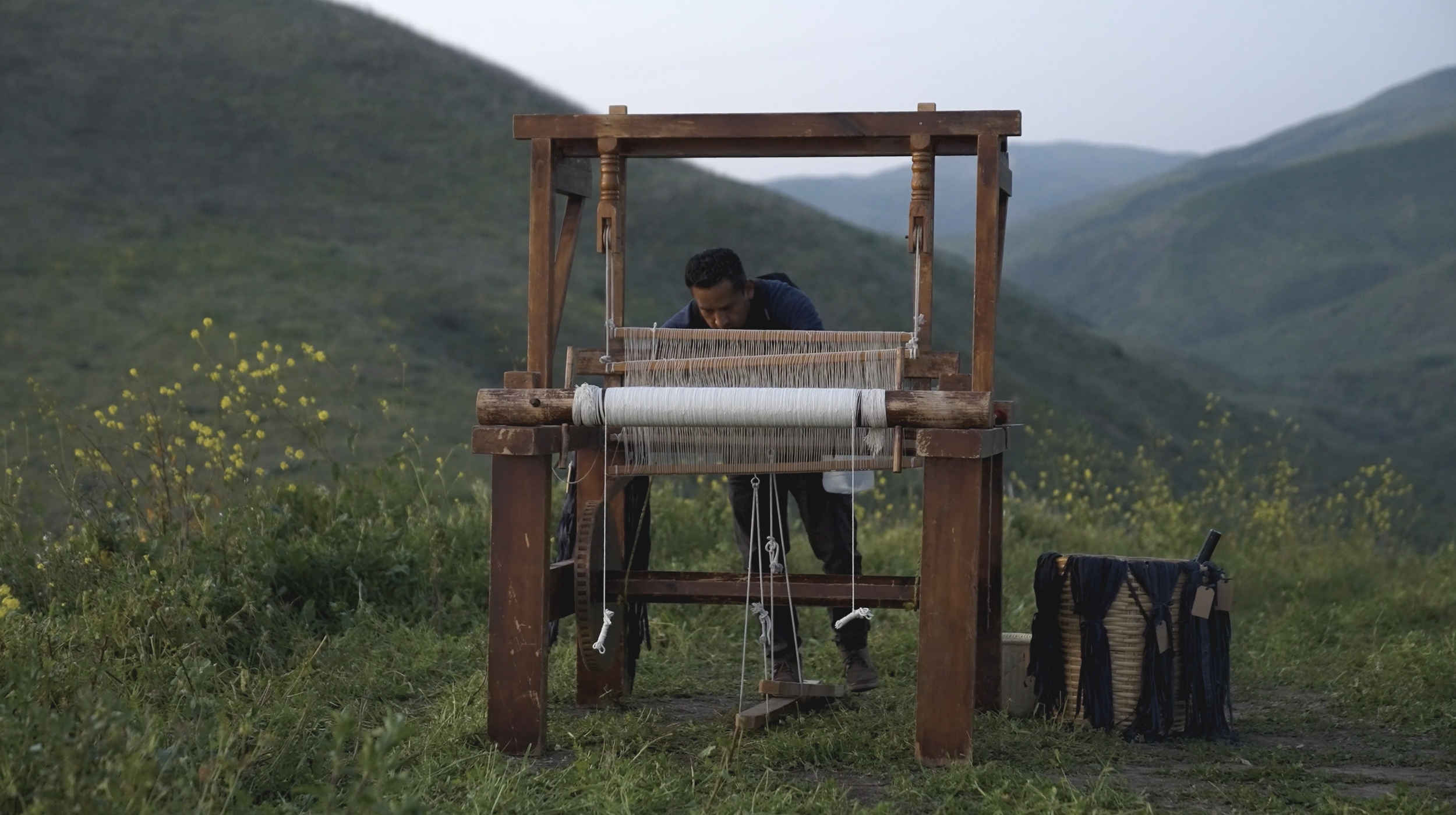A man working on a traditional loom outdoors in a green, mountainous landscape with yellow flowers.