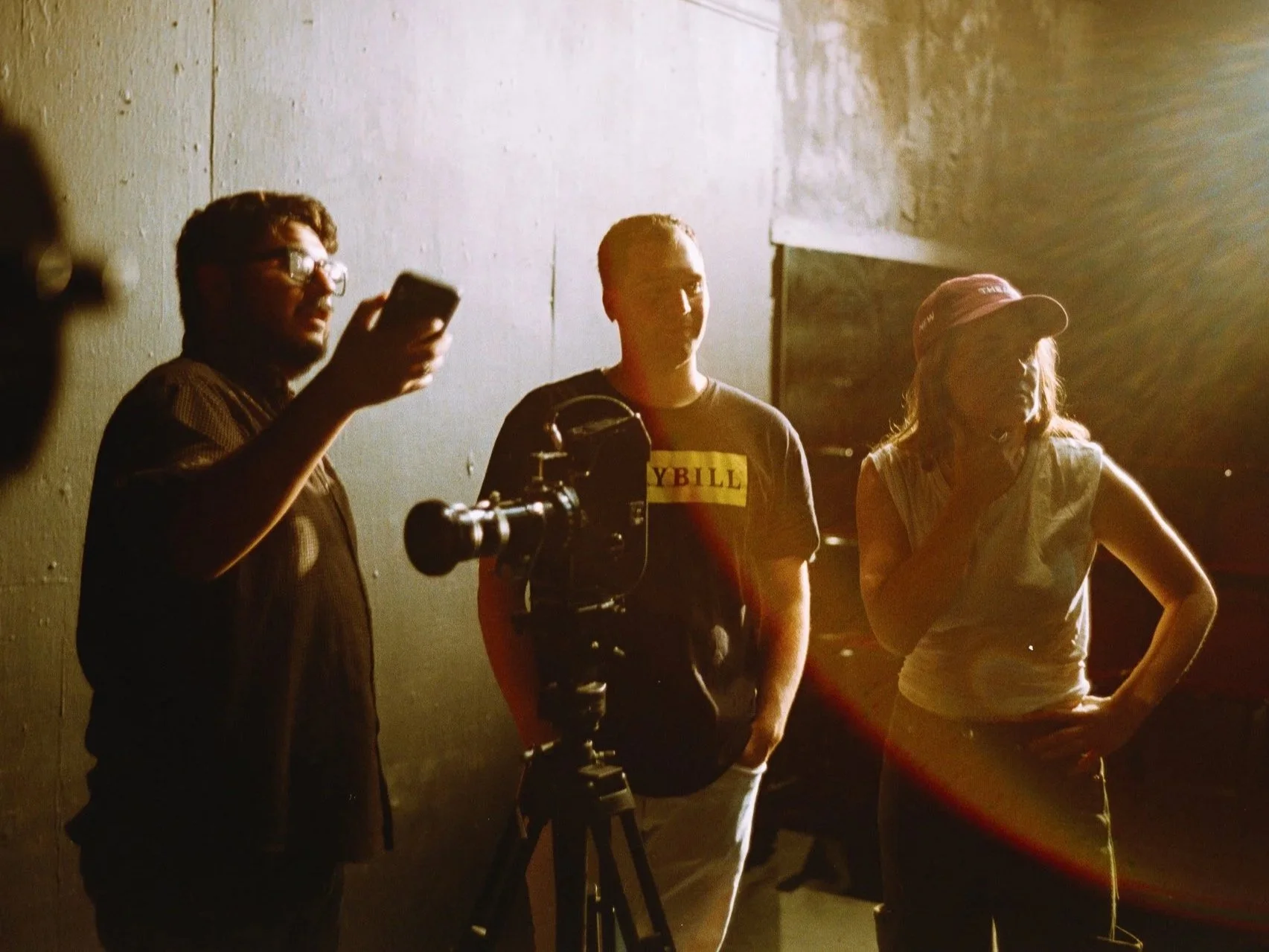 Three people standing together against a concrete wall, with a camera on a tripod in front of them. They are in dim lighting, with sunlight creating a strong backlight.