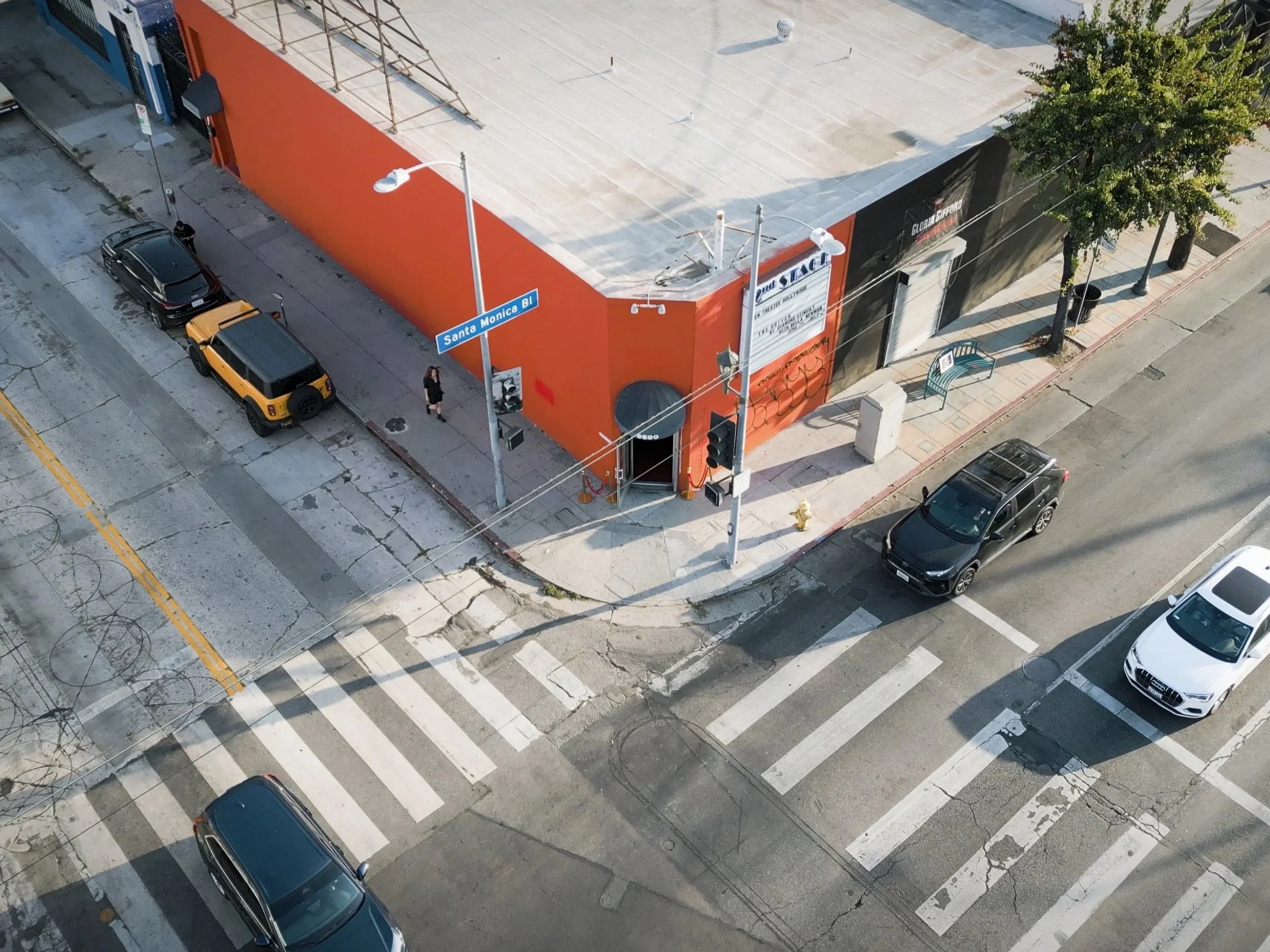 Bird's eye view of a city street corner with cars parked along the curb, a woman walking on the sidewalk, a large orange building with a sign, trees, street lights, utility poles, and a crosswalk.