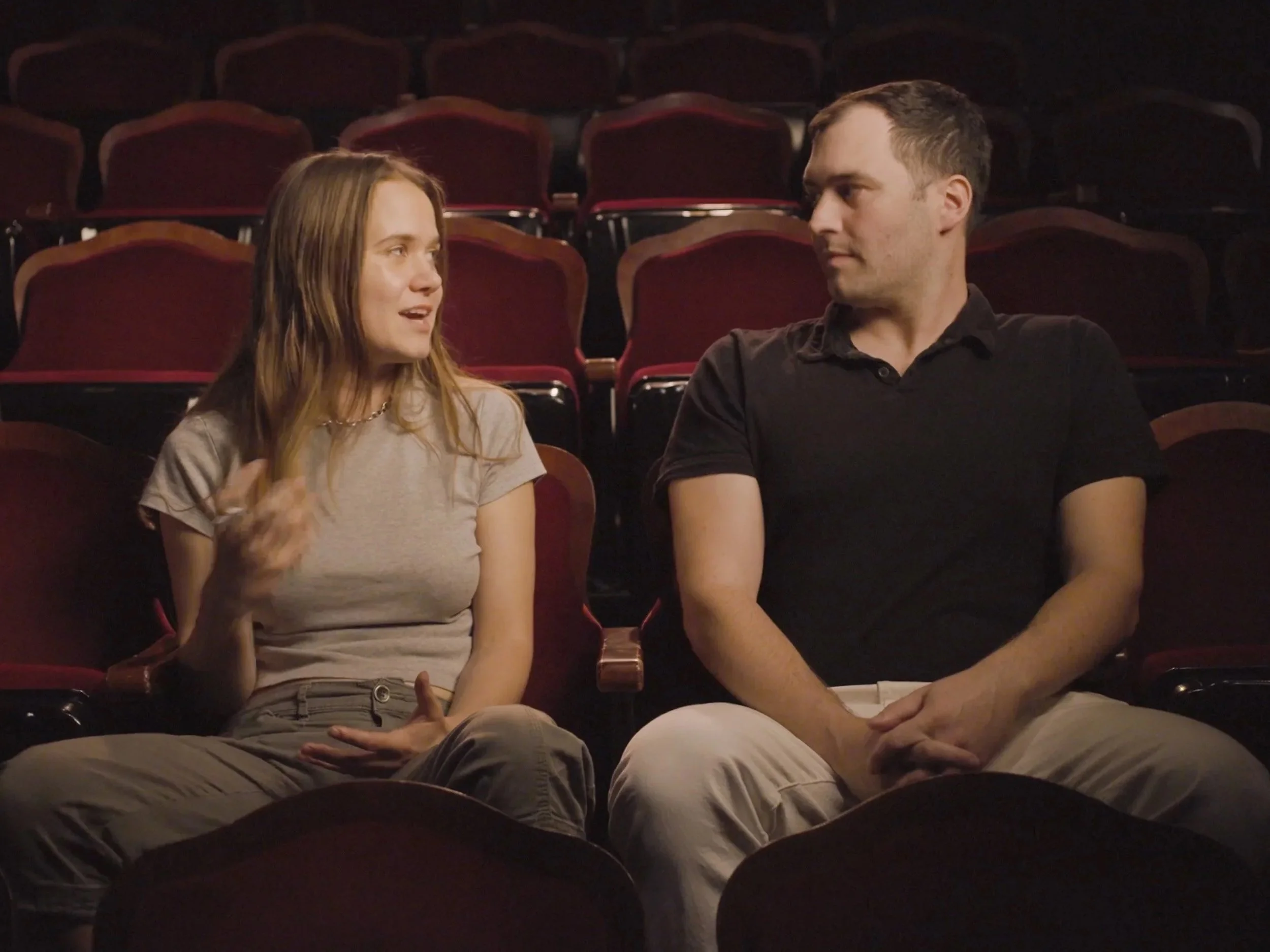 A woman and a man sit in an empty theater, engaged in conversation.