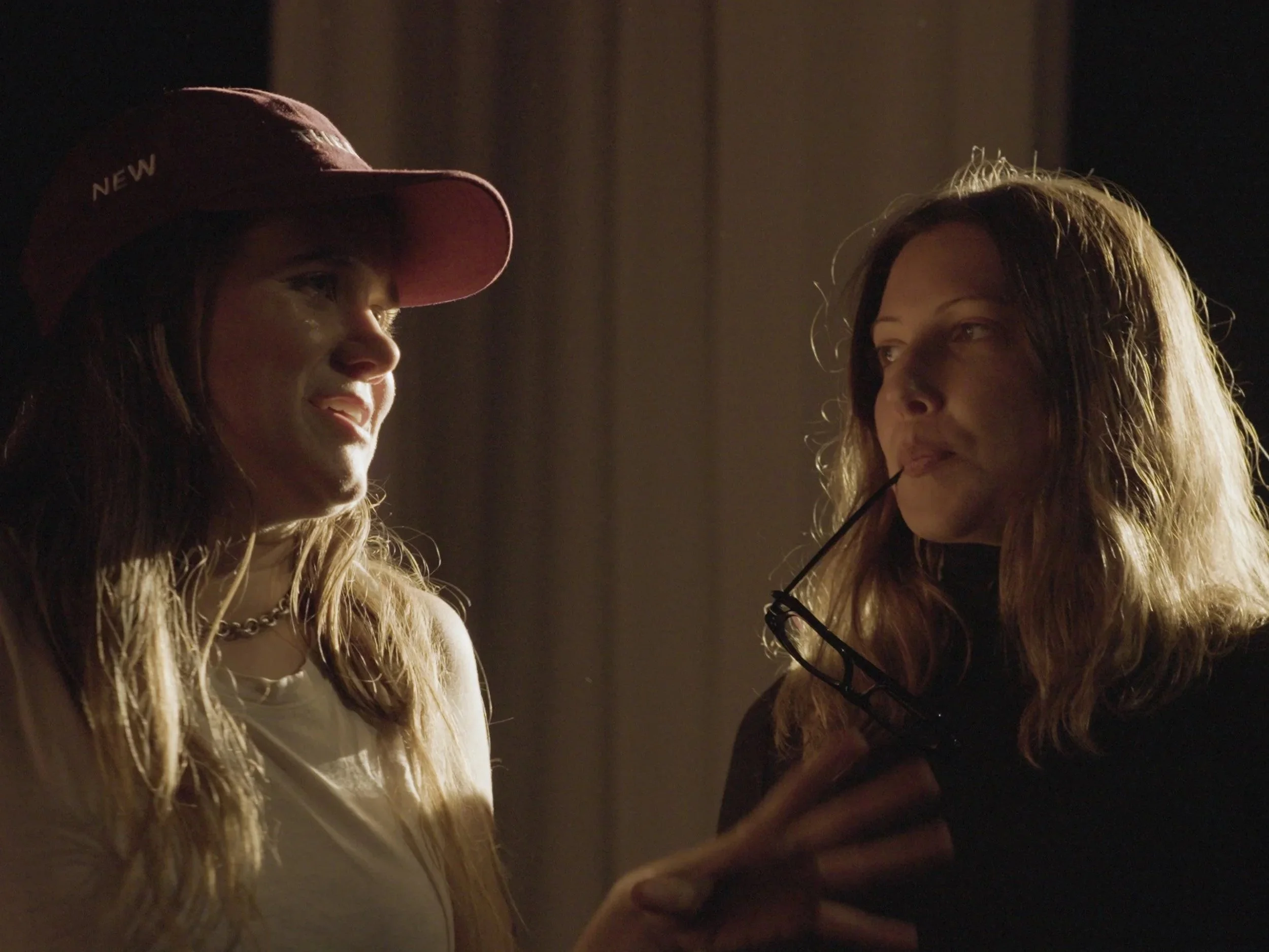 Two women are in a dimly lit room. The woman on the left is wearing a cap with the word "NEW" and is smiling, while the woman on the right has glasses hanging from her mouth and is looking at her.